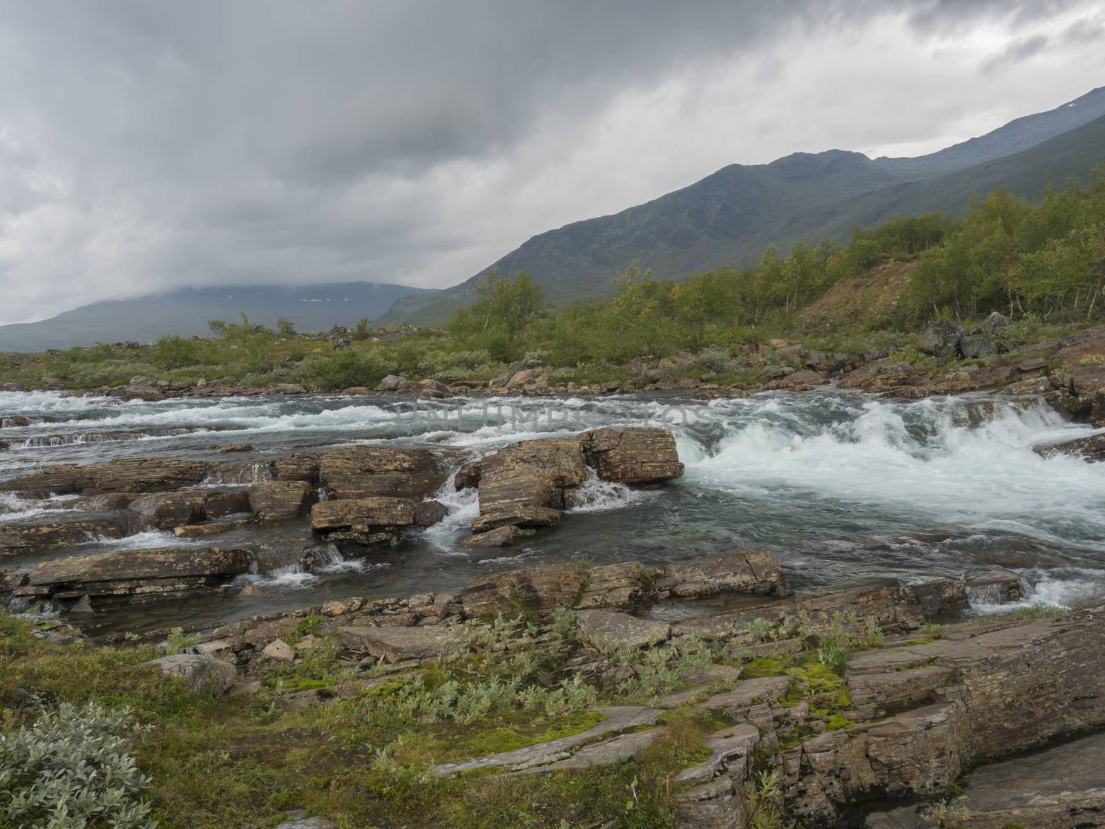 Wild Tjaktjajakka river with waterfall cascade, birch tree forest and mountains. Lapland nature landscape in summer, moody sky