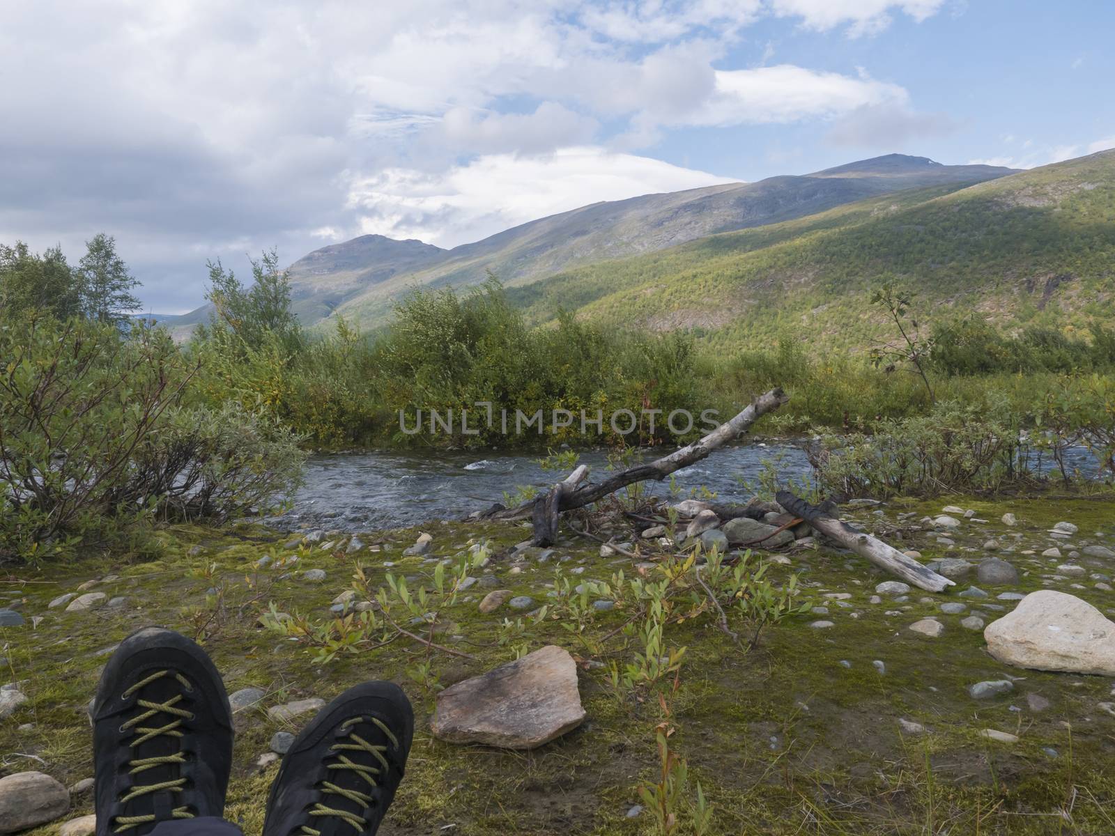 hiking boots with view on wild river with birch tree forest and mountains in golden light. Lapland nature landscape in summer, moody sky