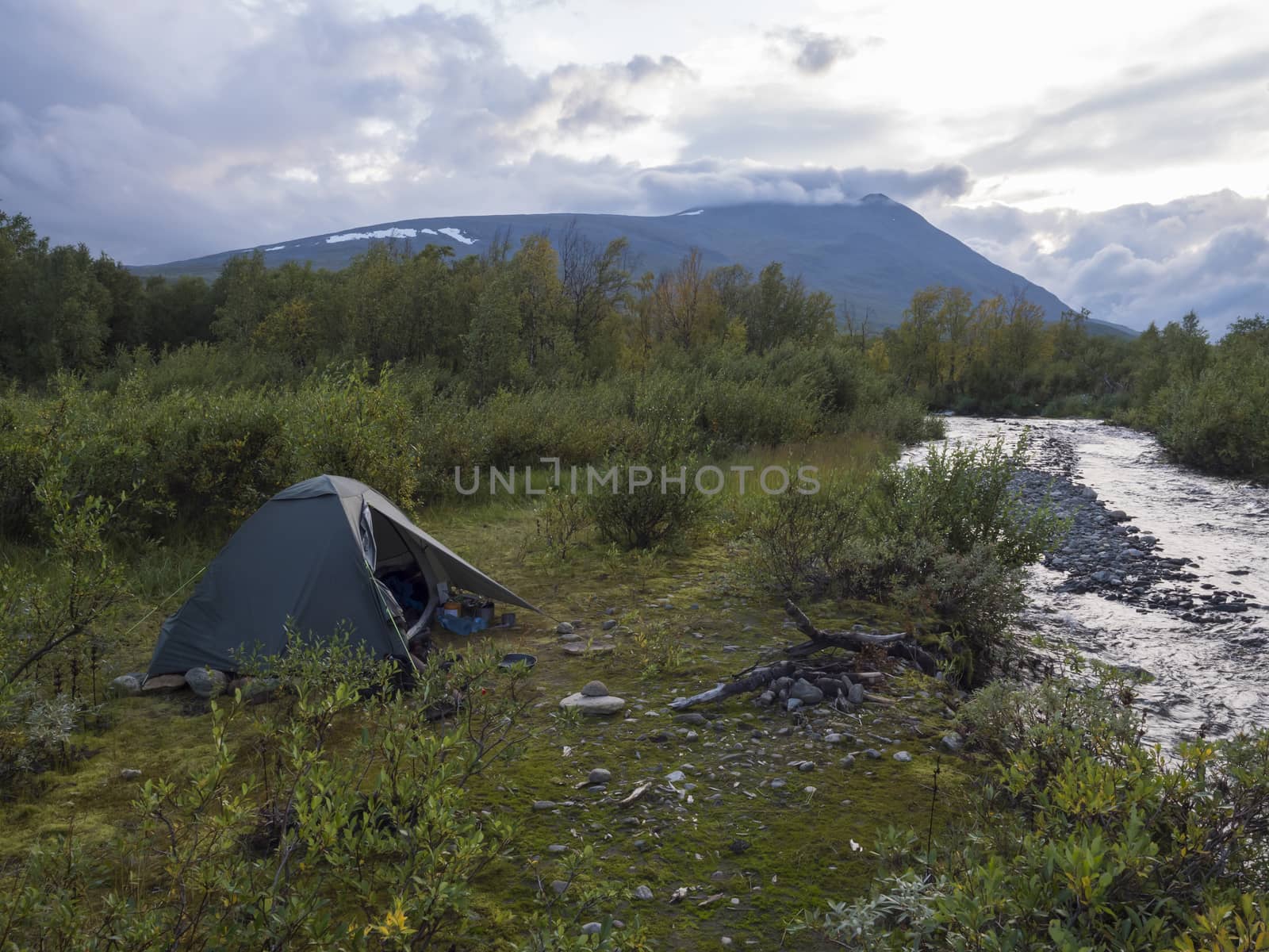 Small green tent in Beautiful wild Lapland nature landscape with blue river, birch trees and mountains. Northern Sweden summer at Kungsleden hiking trail. Blue sky dramatic clouds.