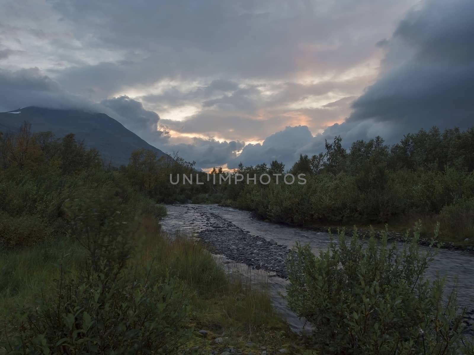 wild river with birch tree forest and mountains in sunset light. Lapland nature landscape in summer, moody sky, pink clouds