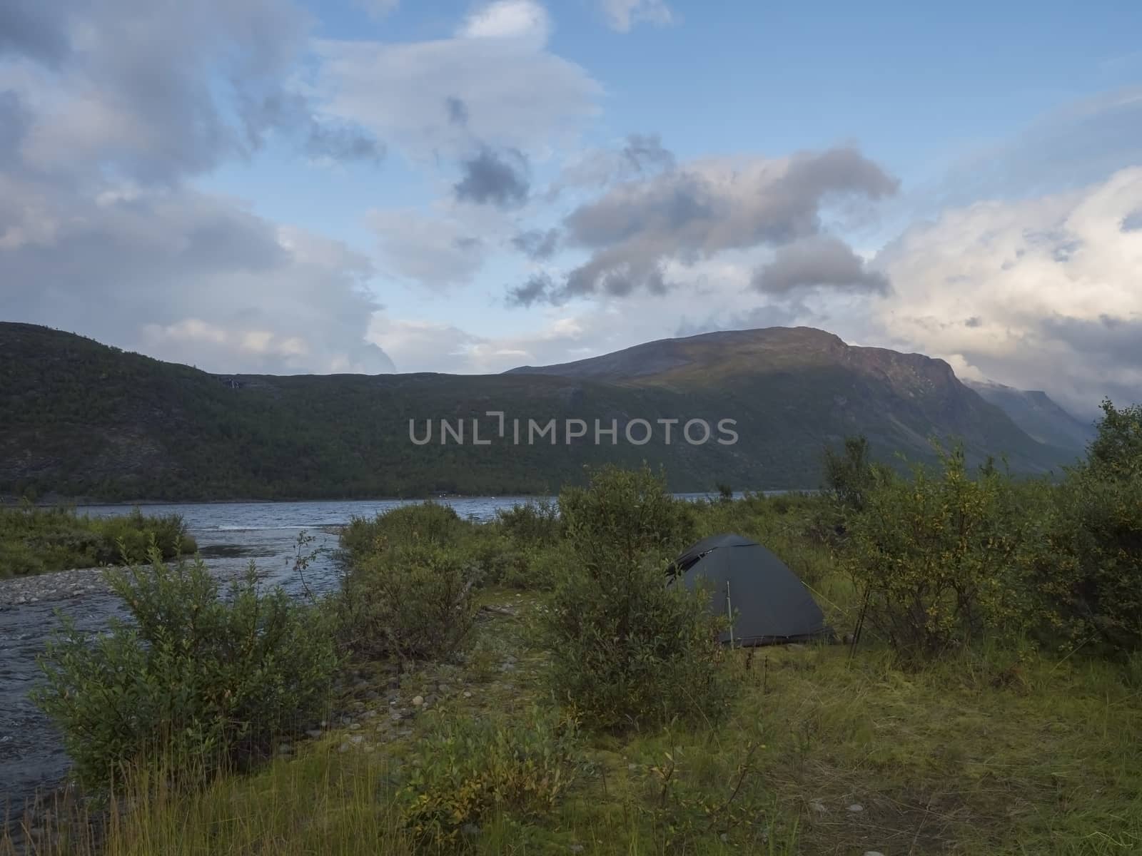 Small green tent in Beautiful wild Lapland nature landscape with blue river, birch trees and mountains. Northern Sweden summer at Kungsleden hiking trail. Blue sky dramatic clouds.