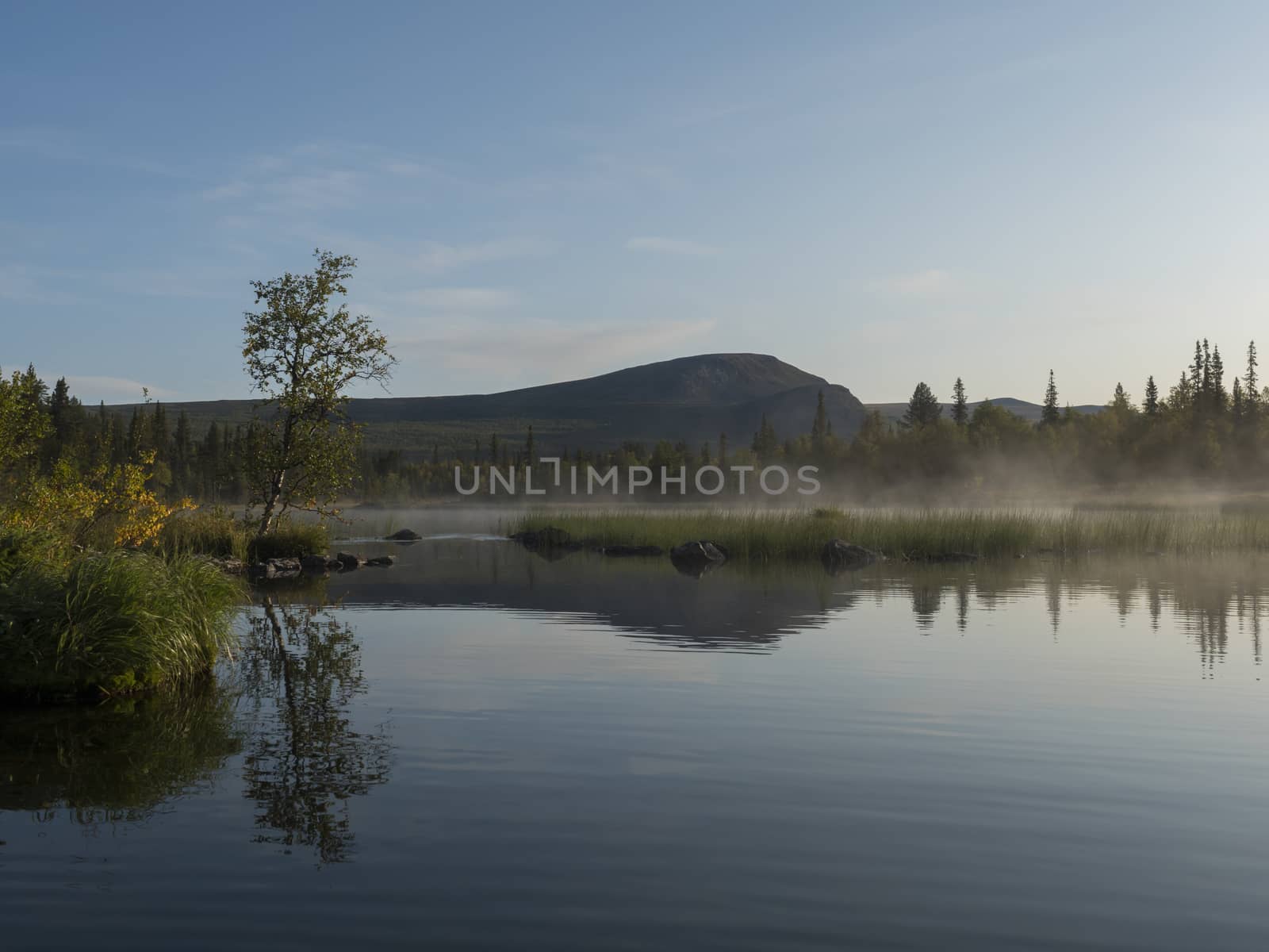 Beautiful morning sunrise over lake Sjabatjakjaure with haze mist in Sweden Lapland nature. Mountains, birch trees, spruce forest, rock boulders and grass. Sky, clouds and clear water