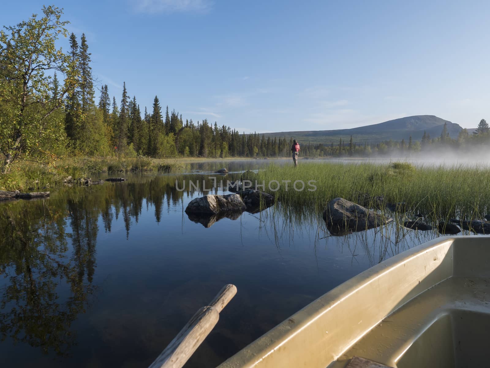 View from rowing boat on Fisherman man at lake Sjabatjakjaure in Beautiful sunny morning haze mist in Sweden Lapland nature. Mountains, birch trees, spruce forest. Blu sky background.