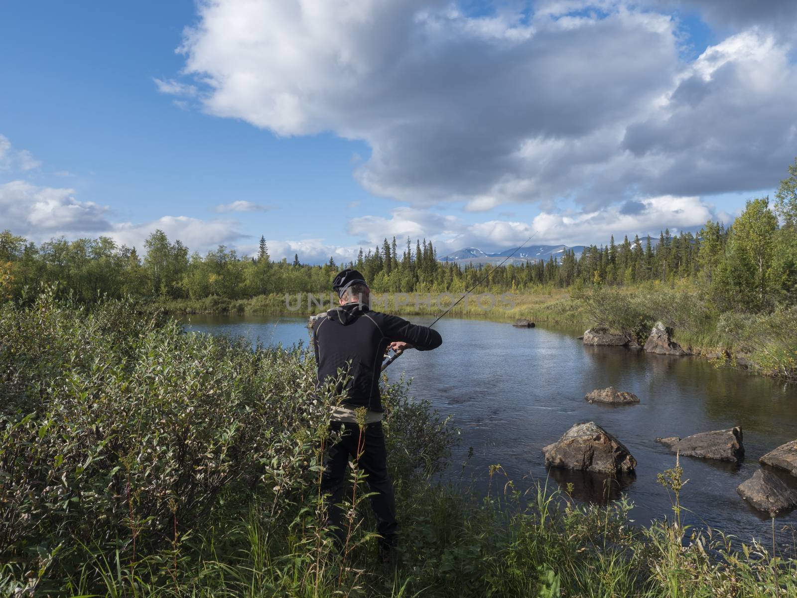 Fisherman man catching fish at beautiful northern landscape with azure blue river Kamajokk, mountains, boulders and spruce tree forest in Kvikkjokk in Swedish Lapland. Summer sunny day, white clouds.