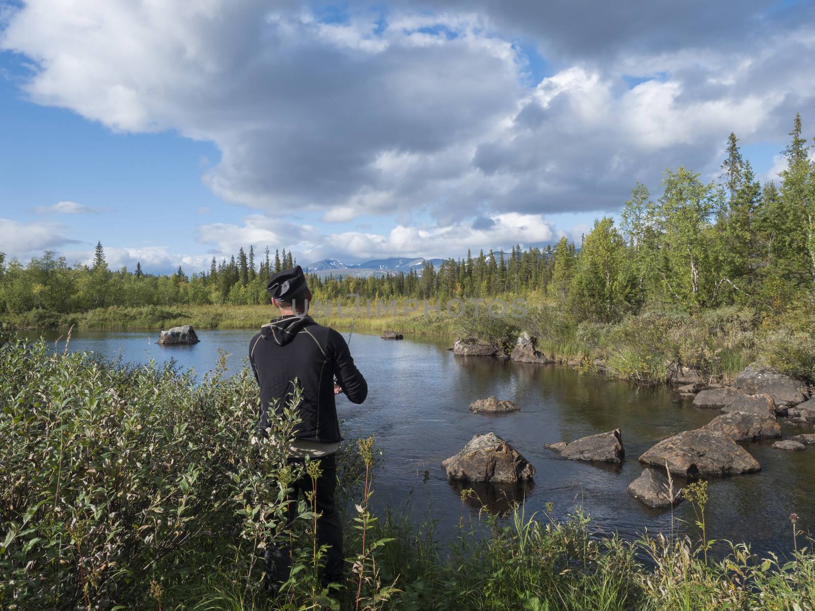 Fisherman man catching fish at beautiful northern landscape with azure blue river Kamajokk, mountains, boulders and spruce tree forest in Kvikkjokk in Swedish Lapland. Summer sunny day, white clouds.