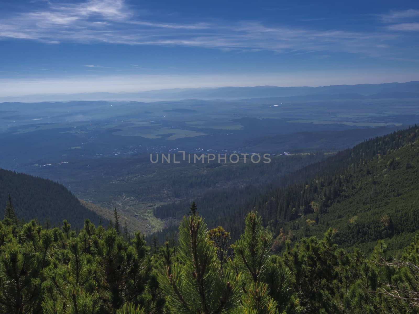 View from tatra mountains trail on valley with Tatranska lomnice and blue misty slopes of hills in the distance. Pine trees and coniferous forest hills, blue sky. Travel background, tatra mountain in summer, Slovakia.
