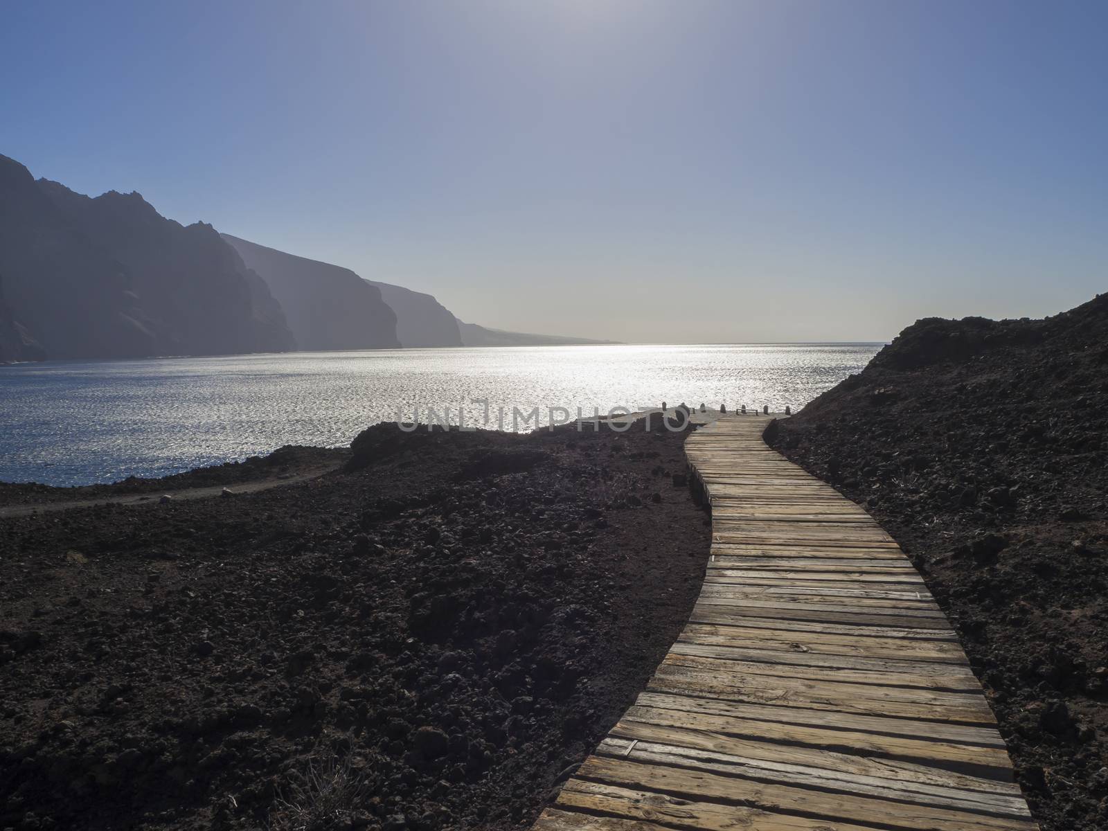 wooden planks path leading to the sea shore with view on hight lava cliffs los gigantes on Punta de Teno cape on west Tenerife canary island with lava rock blue sea and clear summer sky Contre-jour against daylight