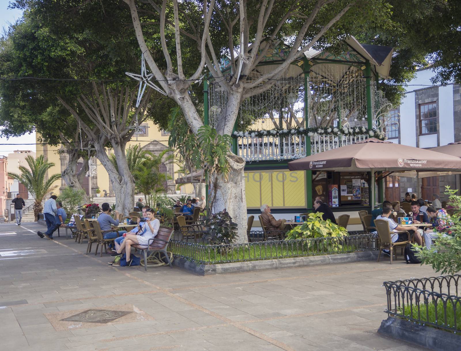 Spain, Canary islands,Garachico, December 19, 2017, group of people and tourist siting on the bench and fast food tables and relaxing in the public park with big trees