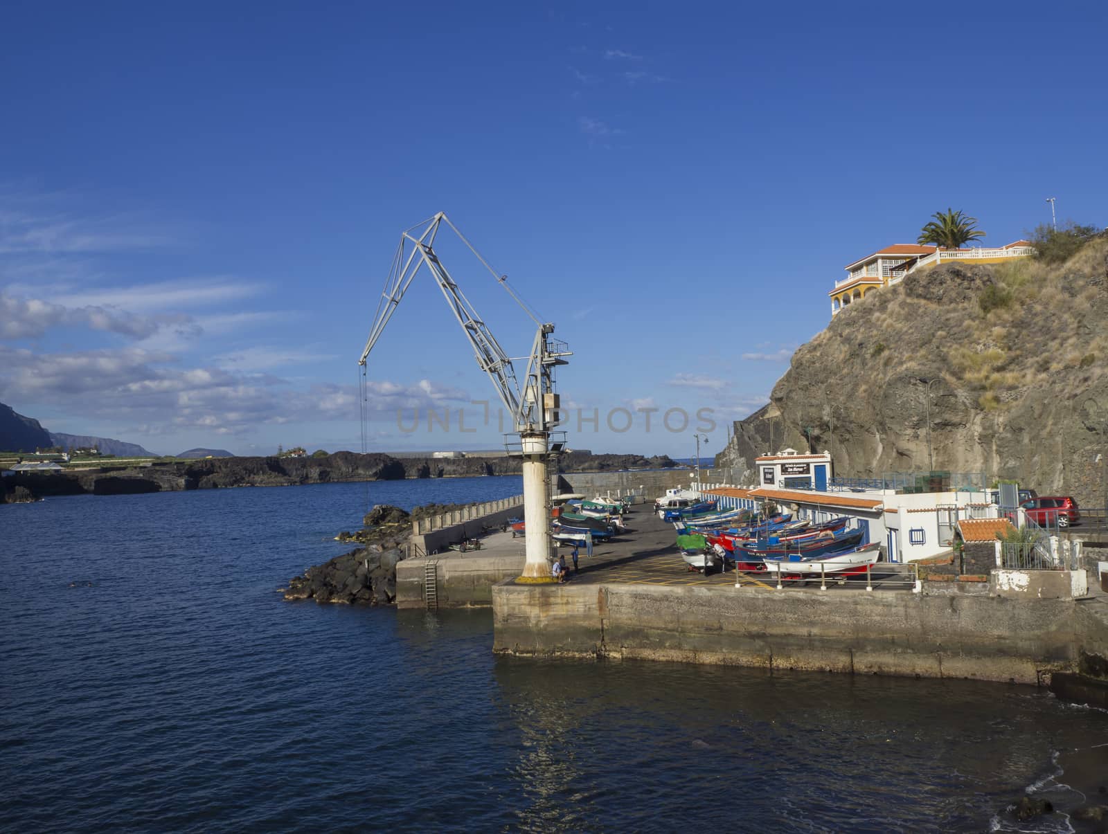 View on fisherman boats dock harbor in San Marcos village on Tenerife with ship crane,  sea, hill and blue sky background