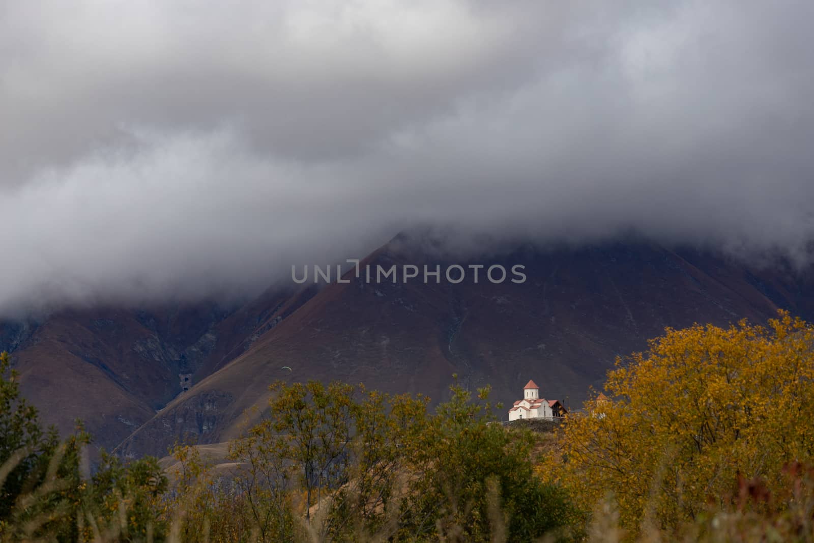 Kazbegi region of Georgia, snow capped mountains and rugged terrain. White Orthodox church with red roofs near Almasiani Georgia with mountains and dark clouds in the background, very atmospheric and darkHigh quality photo