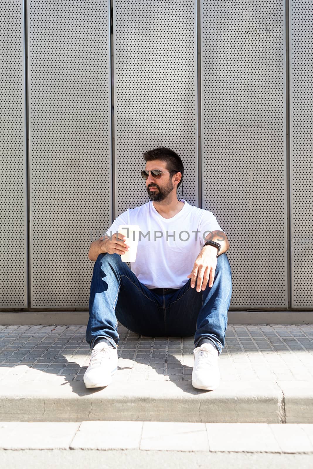 Bearded guy with sunglasses sitting on ground while holding a take away coffee