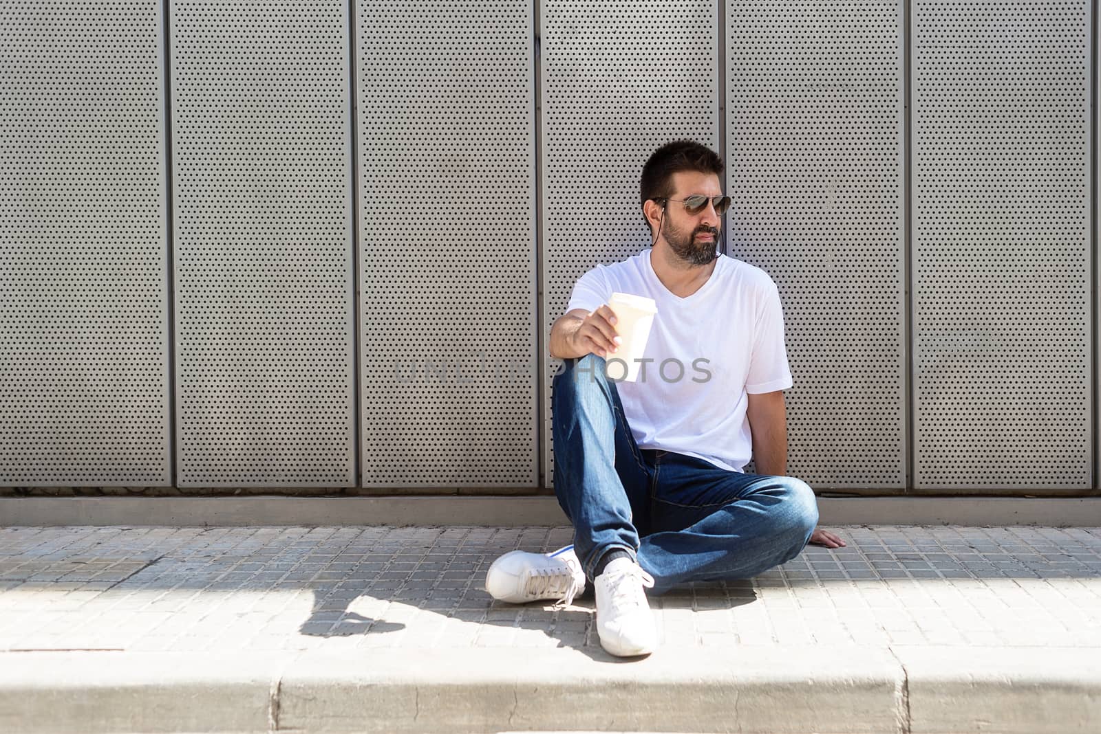 Bearded guy with sunglasses sitting on ground while holding a take away coffee