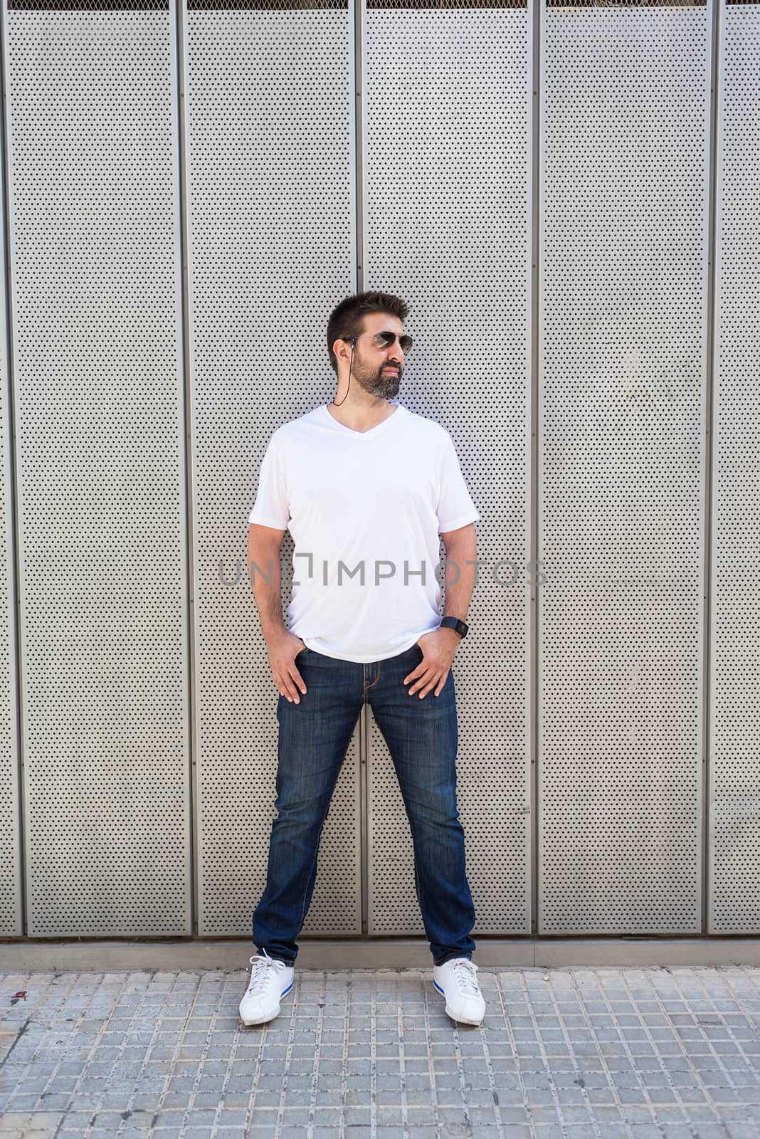 Bearded guy in sunglasses leaning on metallic wall with hands on pocket while looking away