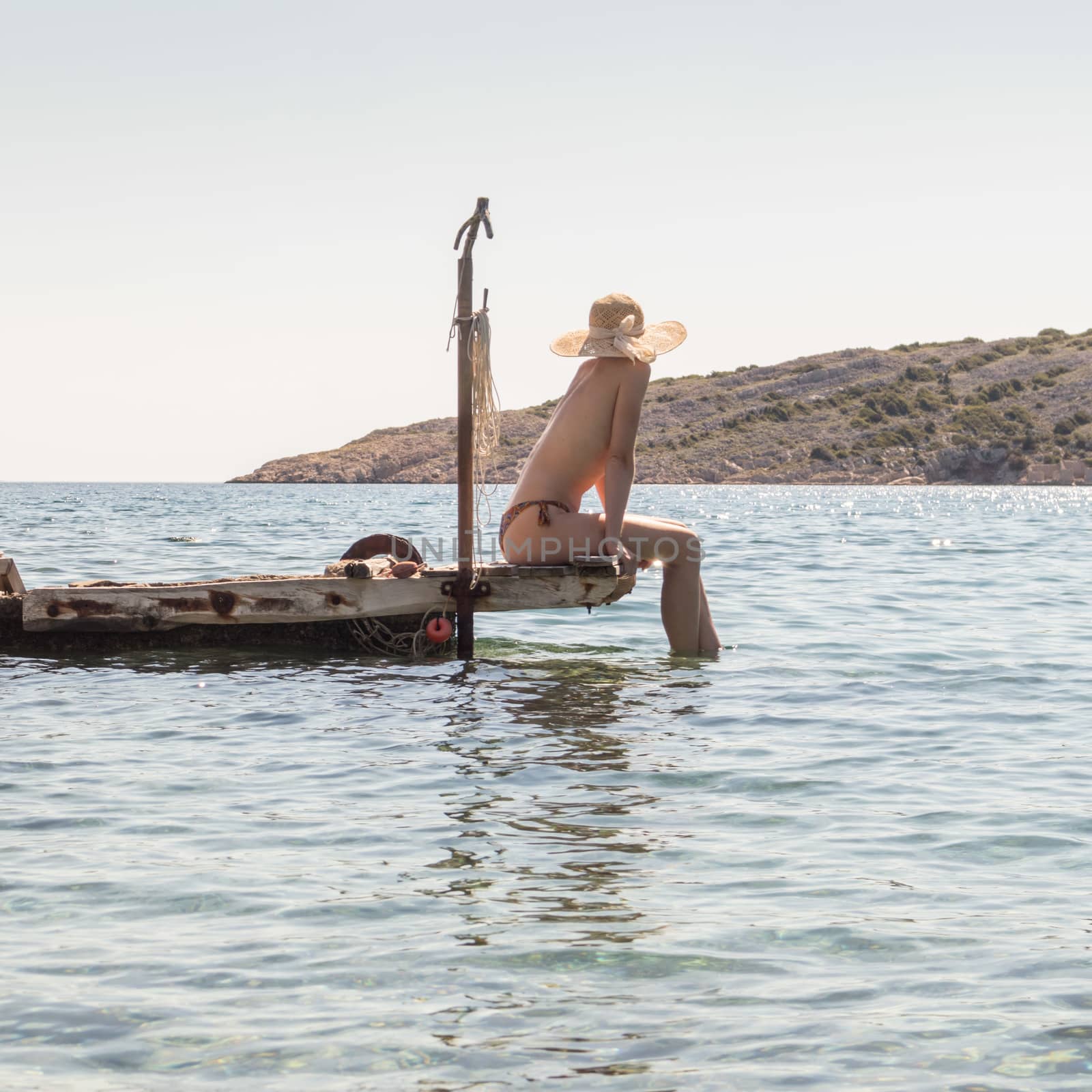 View of unrecognizable woman wearing big summer sun hat tanning topless and relaxing on old wooden pier in remote calm cove of Adriatic sea, Croatia by kasto