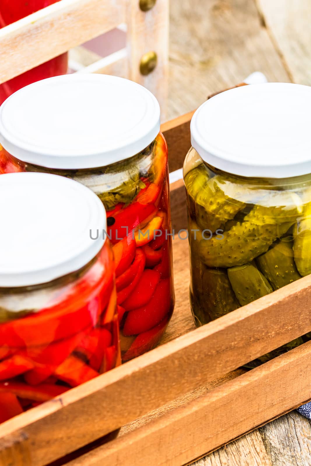 Wooden crate with glass jars with pickled red bell peppers and pickled cucumbers (pickles) isolated. Jars with variety of pickled vegetables. Preserved food concept in a rustic composition.