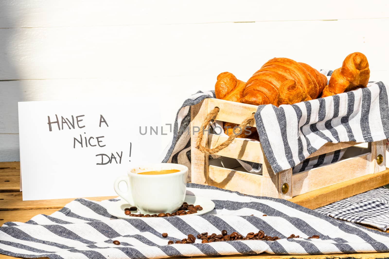 Coffee cup and buttered fresh French croissant on wooden crate. Food and breakfast concept. Morning message “have a nice day” on white board