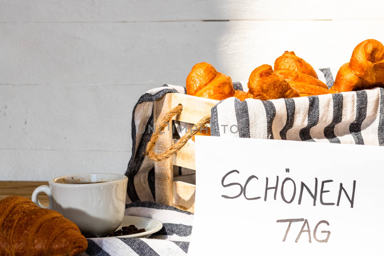 Coffee cup and buttered fresh French croissant on wooden crate. Food and breakfast concept. Morning message in German meaning “have a nice day” on white board