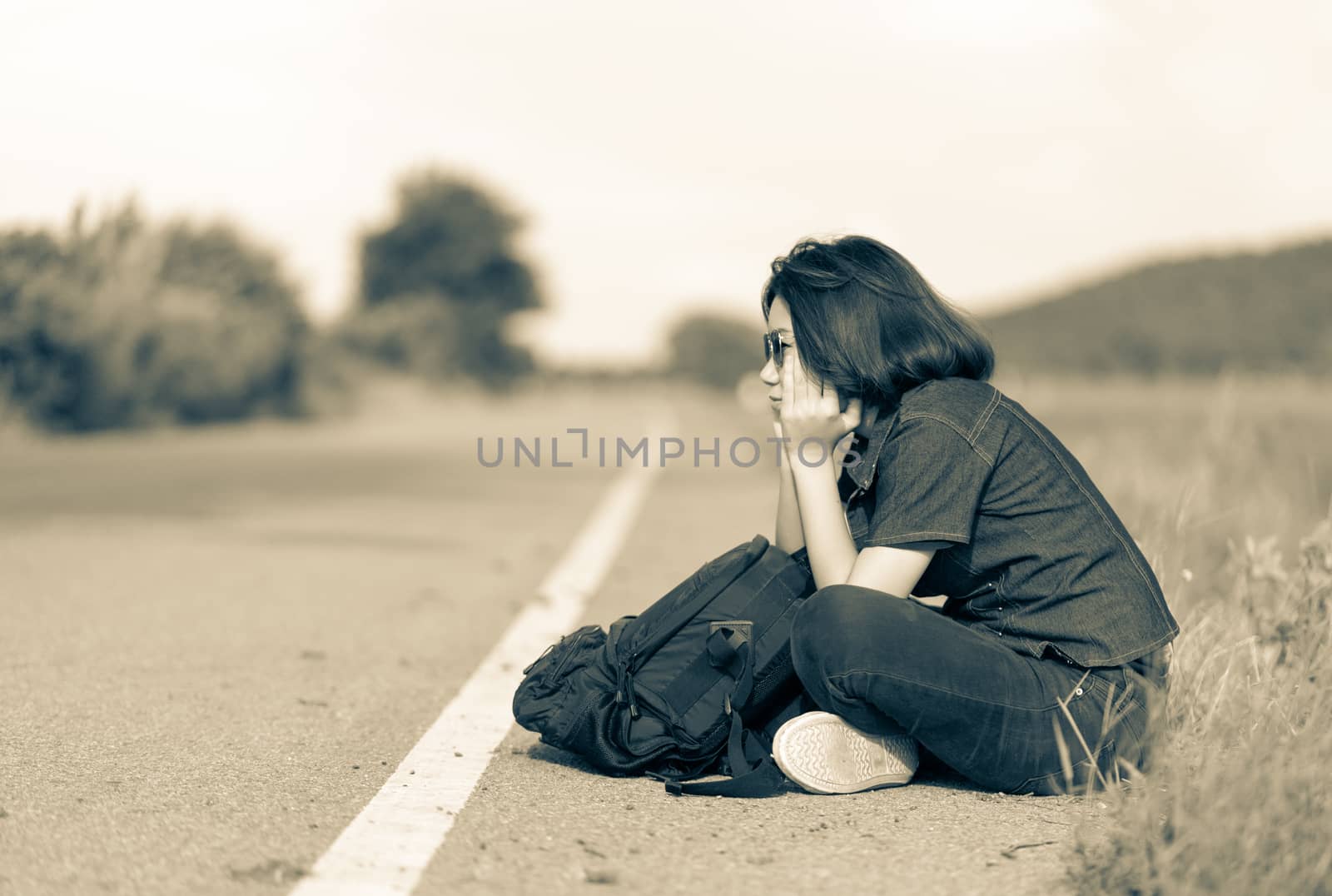 Young asian woman short hair and wearing sunglasses sit with backpack hitchhiking along a road wait for help in country road Thailand