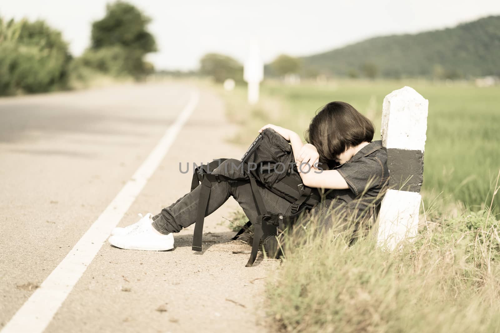 Young asian woman short hair and wearing sunglasses sit with backpack hitchhiking along a road wait for help in countryside Thailand