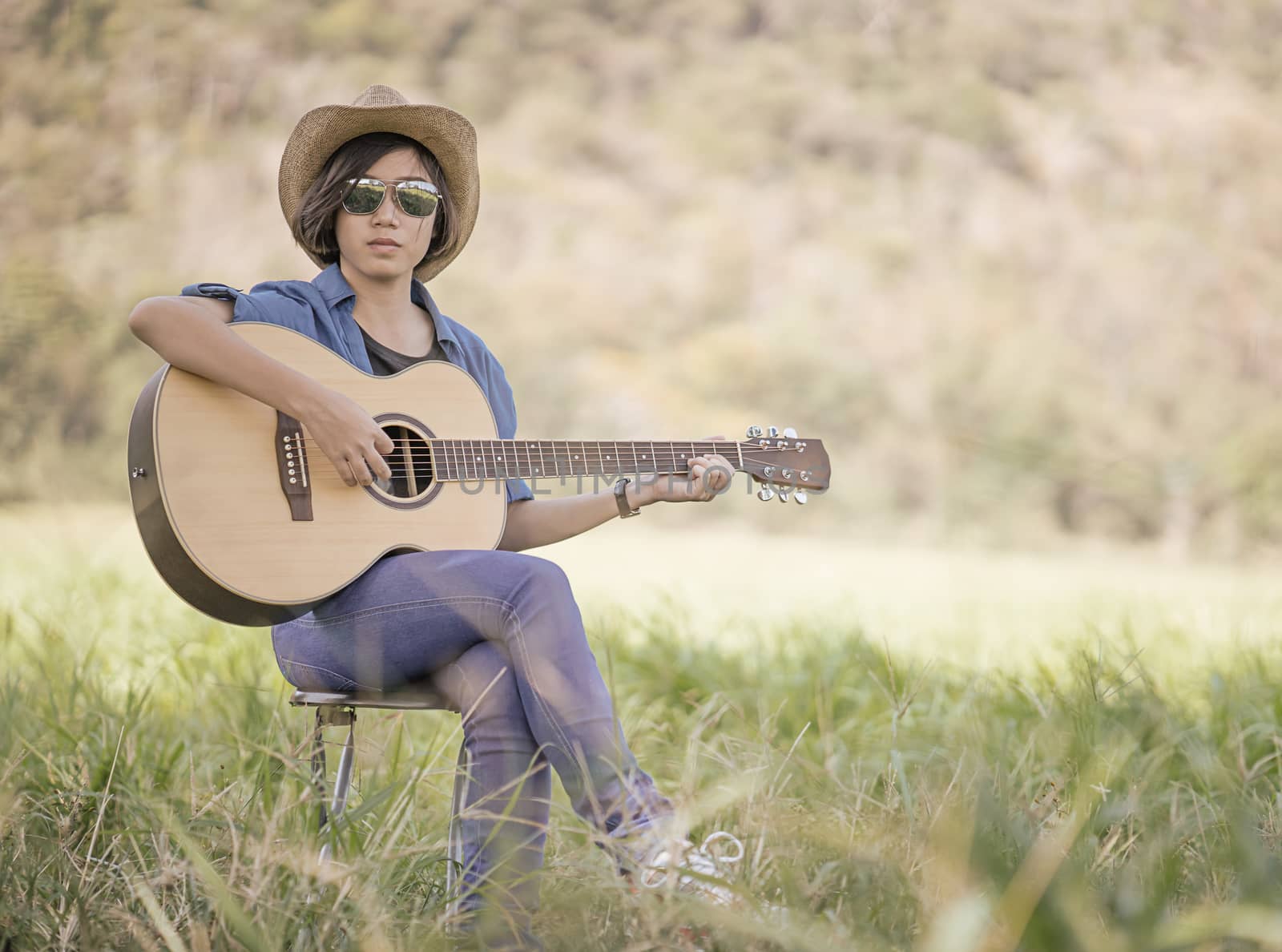 Young asian women short hair wear hat and sunglasses sit playing guitar in grass field countryside Thailand