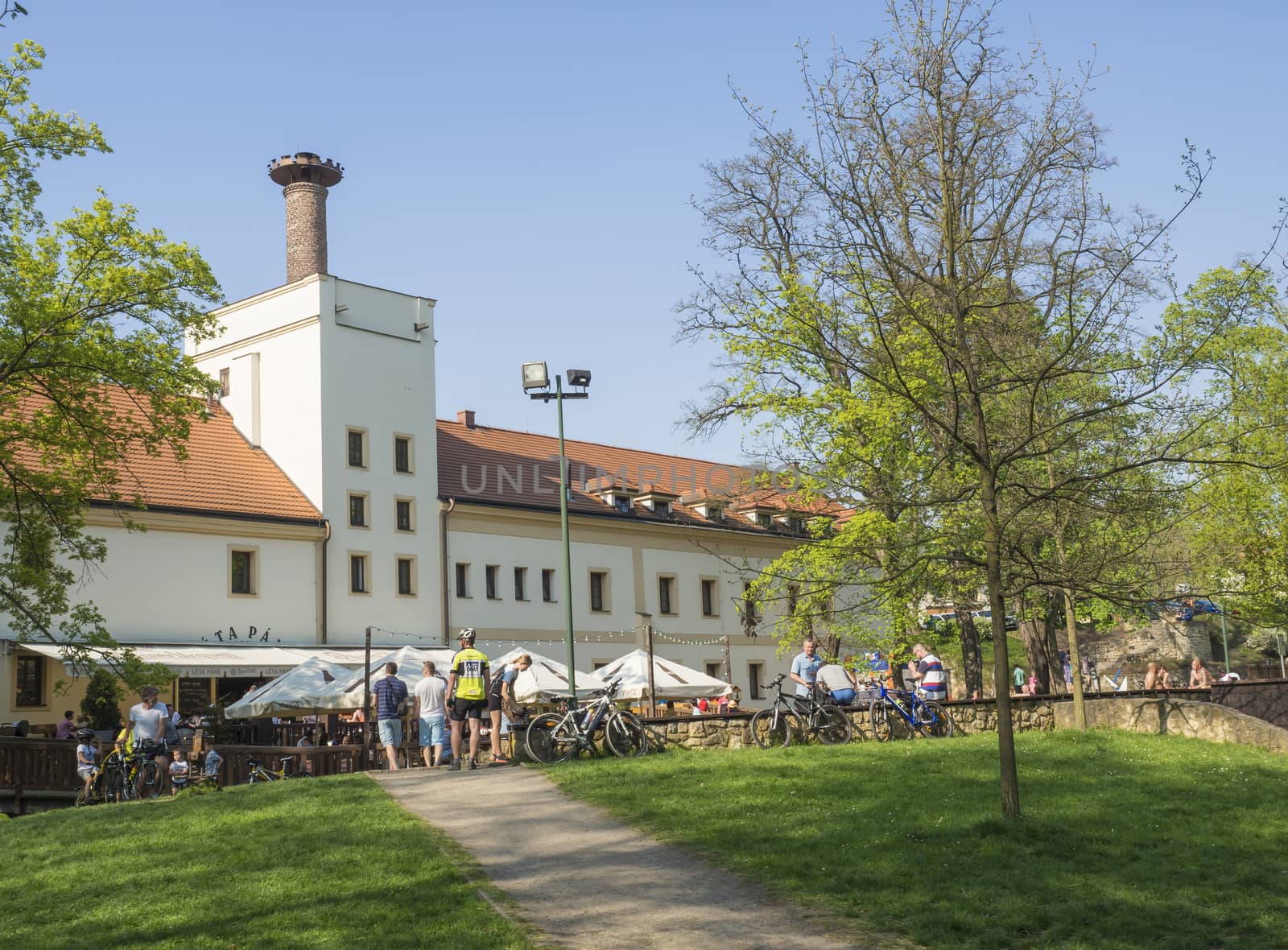 Czech Republic, Prague, Dolni Pocernice, April 21, 2018: old brewery restaurant Pansky dvur Dolni Pocernice with group of people and cyclist relaxing on garden tables, trees, blue sky background, early spring day