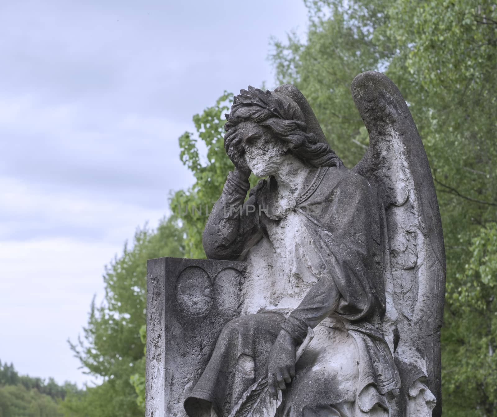 old stone baroque statue of sad greving angel holding head in hand, green tree and blue sky background