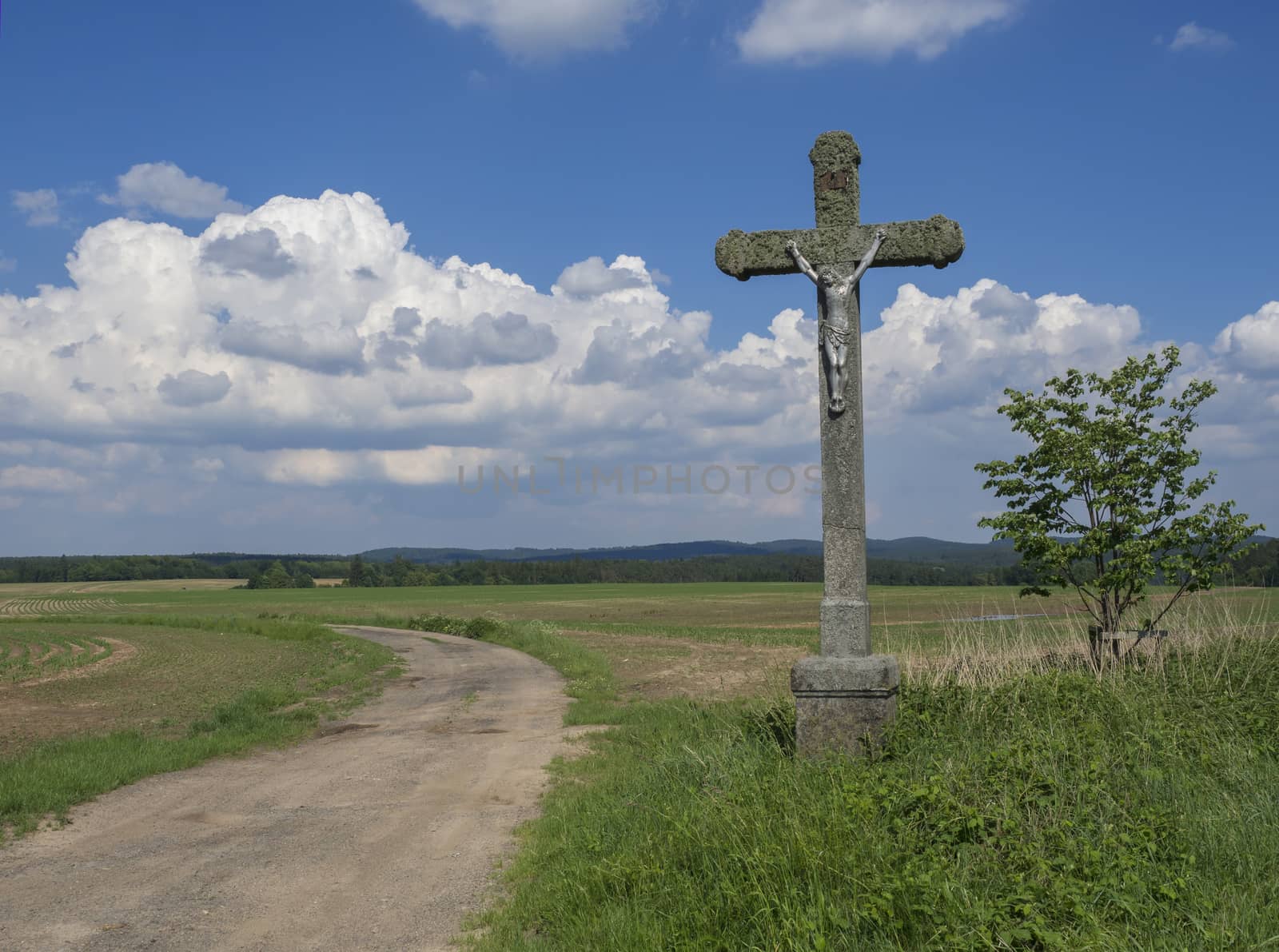 idyllic rural landscape with old stone cross with Jesus Christ statue, crucifix, dirty road, green fields, hills, tree and blue sky white fluffy clouds