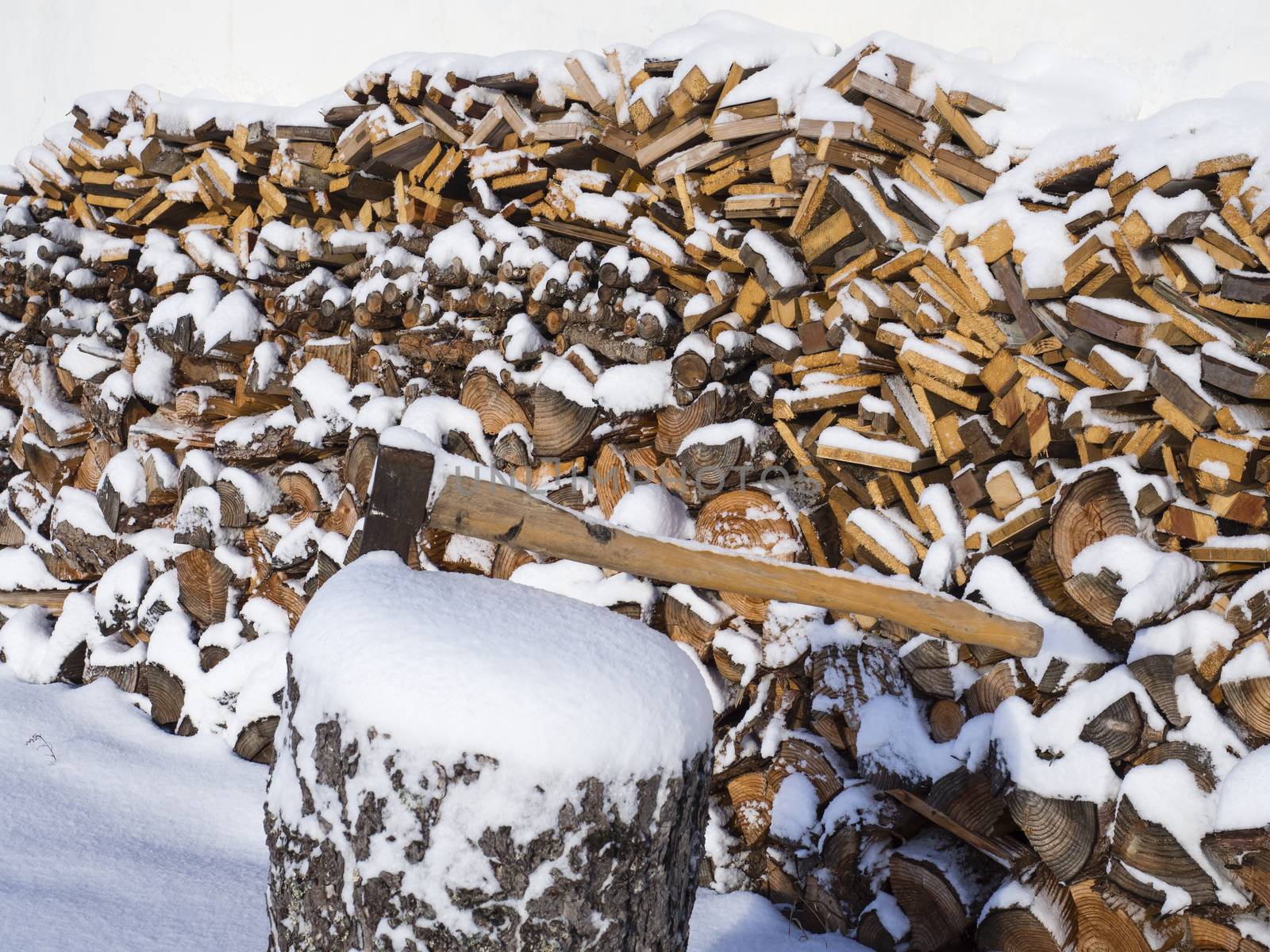 close up axe attached to the trunk of the tree on the background of piled chopped firewood covered with snow.