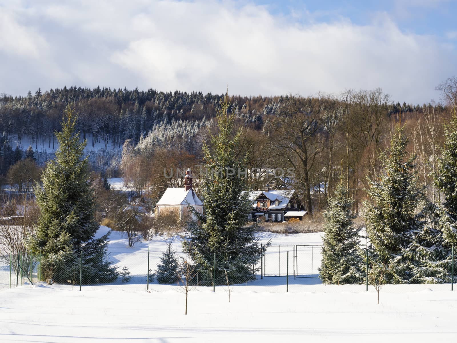 winter view on village travnik with chapel, timbered cottage and trees, snow covered rural landscape with in luzicke hory mountain, sunny day.