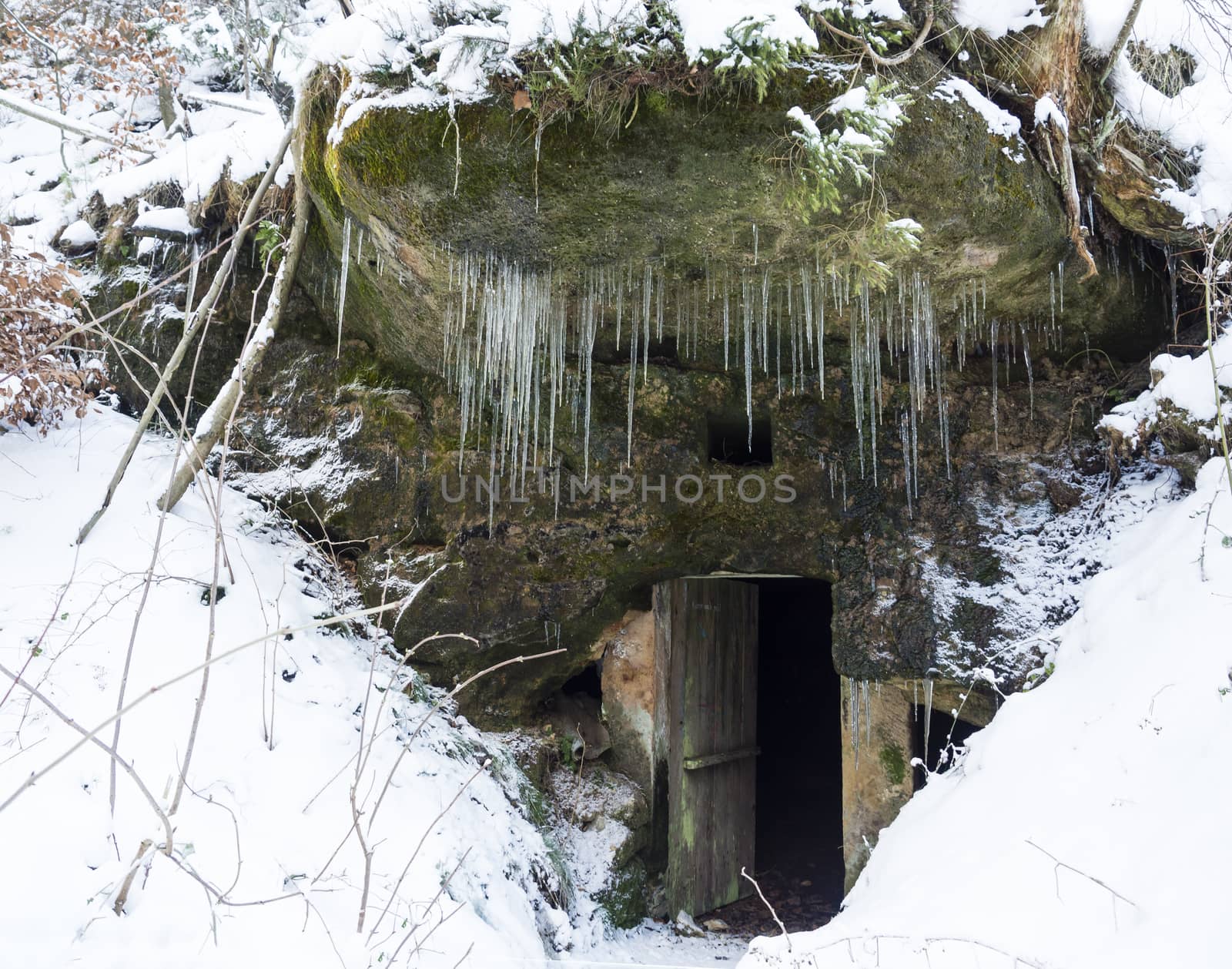 Open dark cellar door in sandstone rock cave with moss and lichens and long hanging icicles, white snow background. by Henkeova