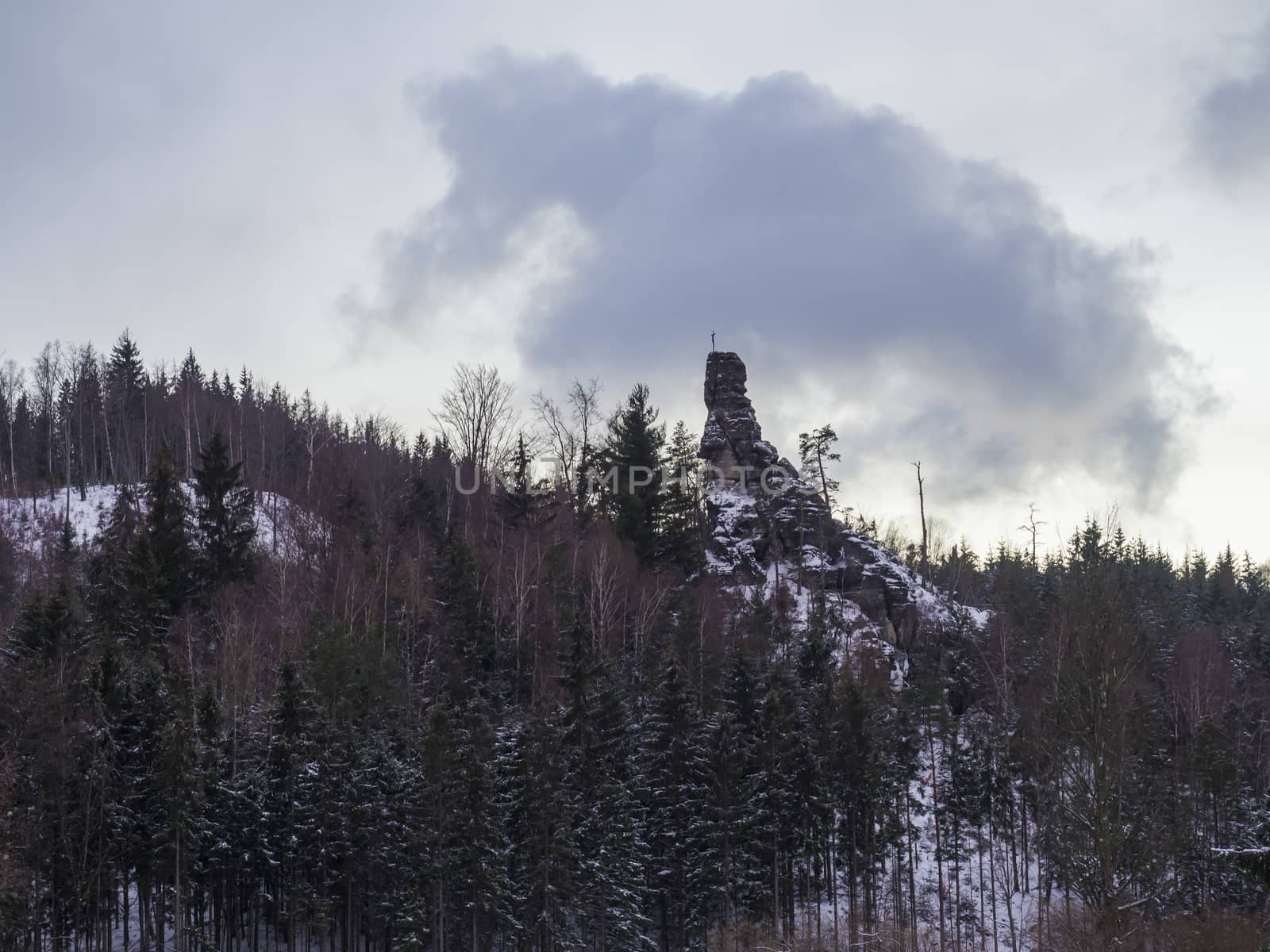 tall sandstone rock with cross and crucifix in snow covered forest landscape with snowy spruce trees and hill, idyllic winter landscape in dusk, blue hour.