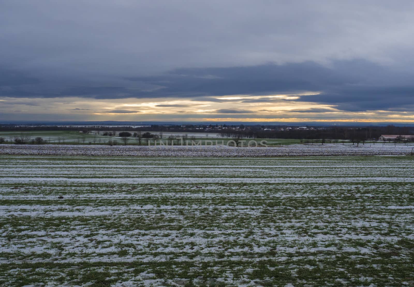 Winter rural landscape with green snow covered fields, bare trees and village houses at orange sunset light. central Bohemian region. Czech Republic. by Henkeova