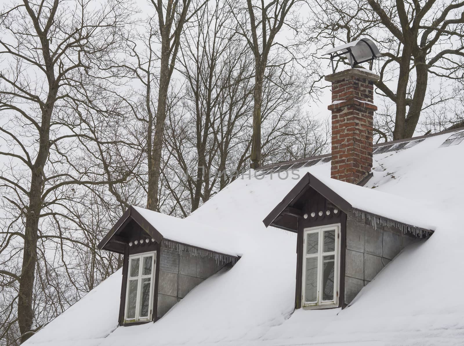 snow covered roof with brick chimney and bay bow window with icicles and bare tree background