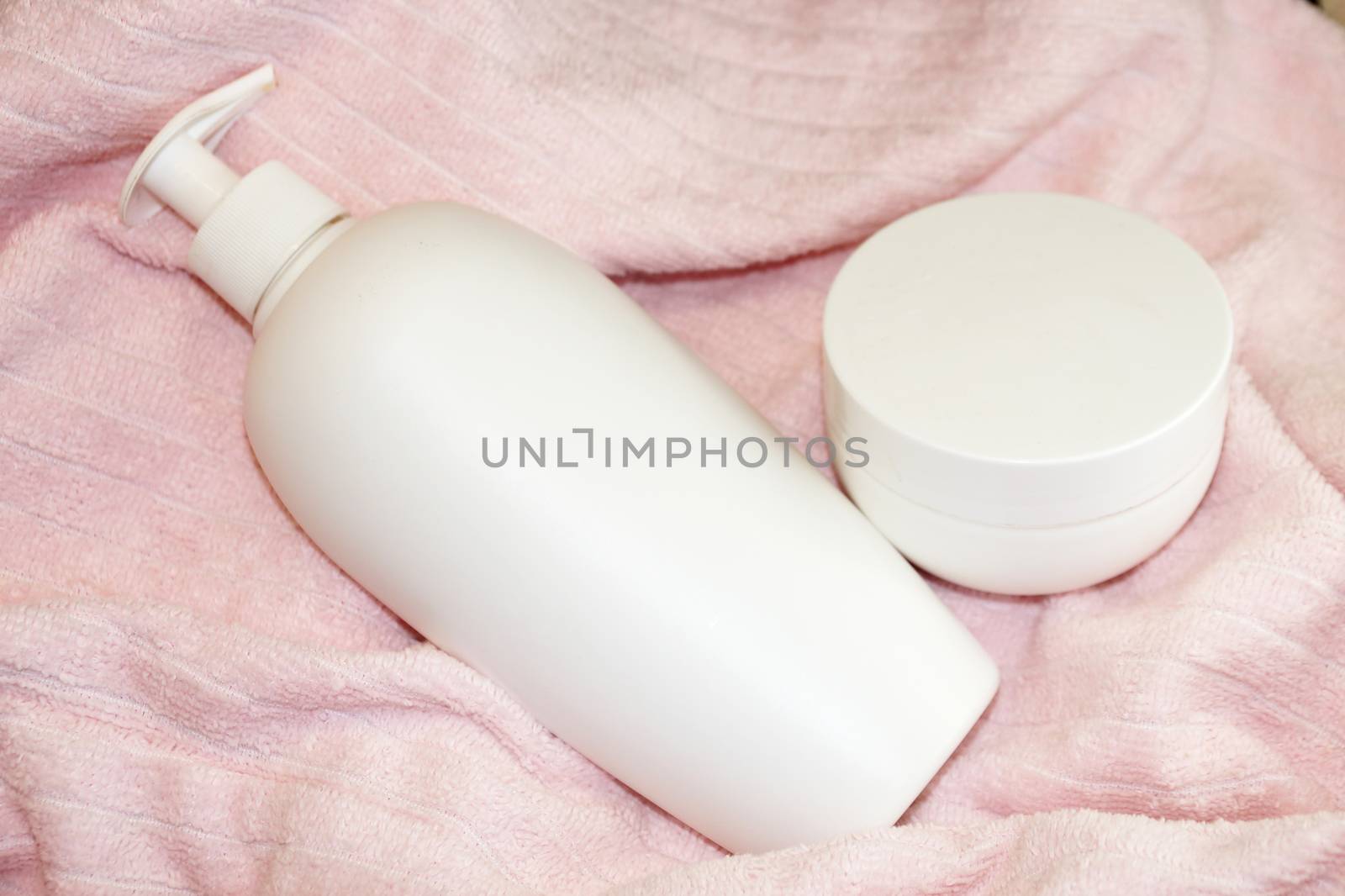 white cosmetic bottle and cream jar on pink terry cloth close-up by Annado