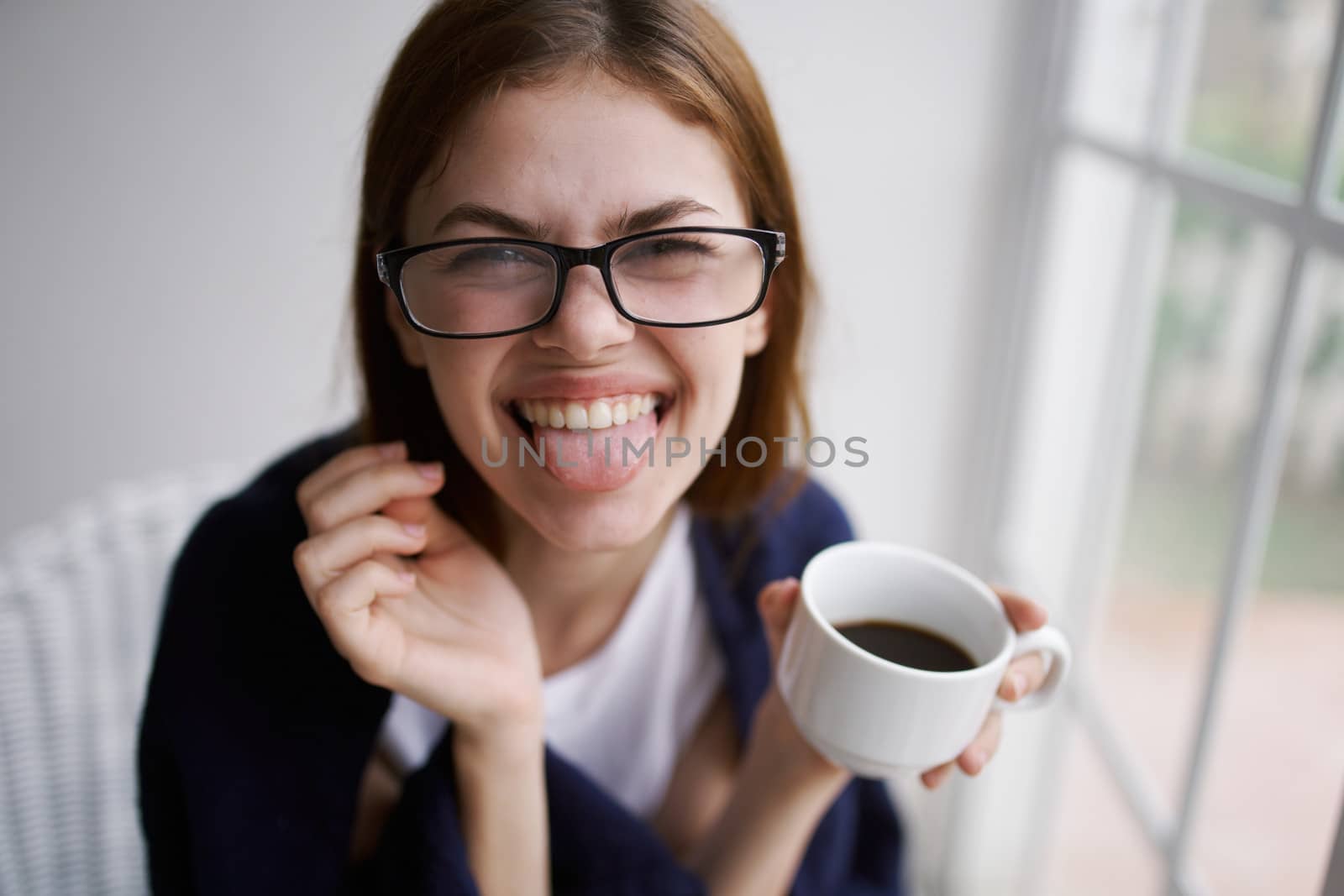 Cheerful woman with a cup of coffee shows tongue to the camera and glasses by SHOTPRIME