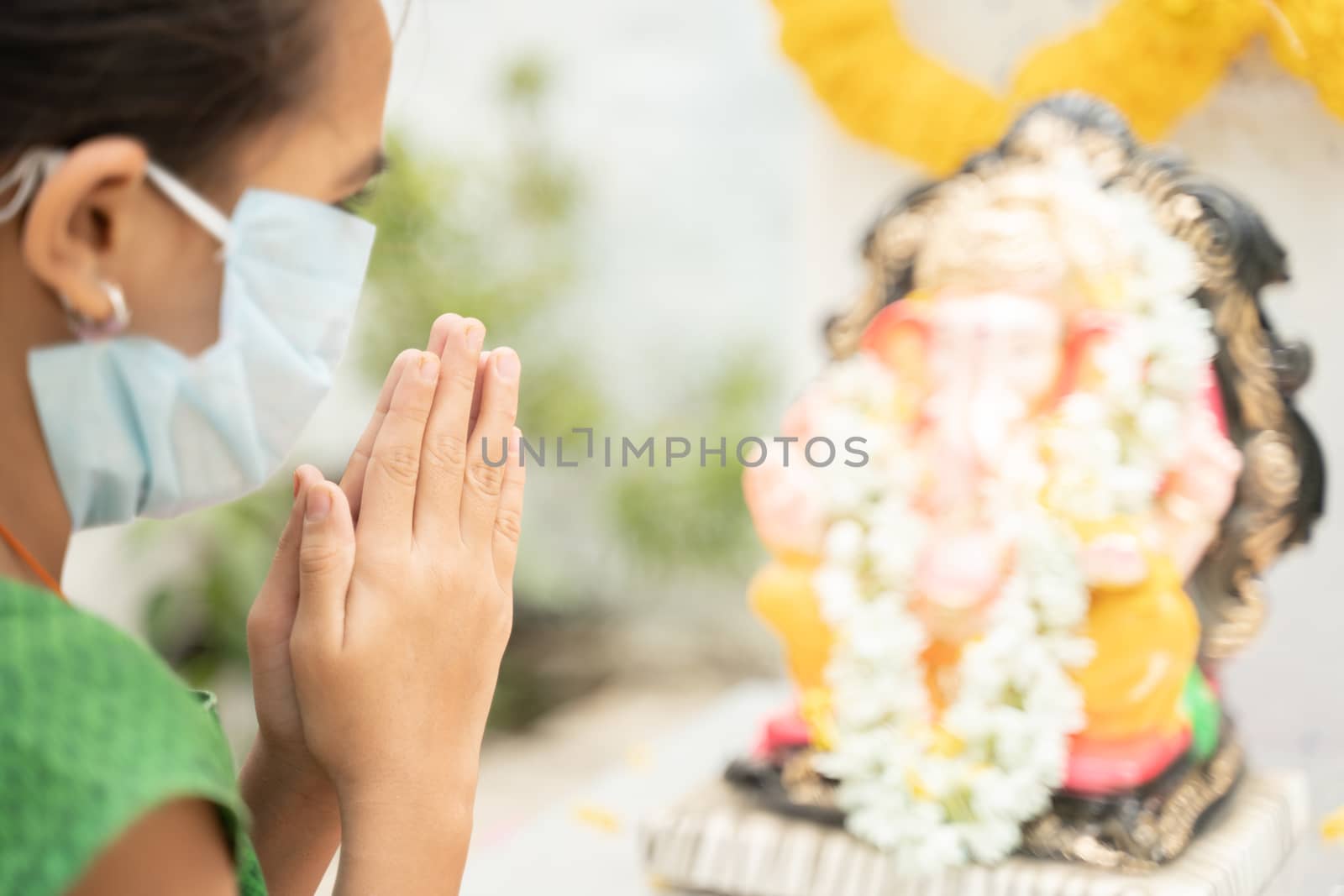 Girl Kid in medical mask praying by closing eyes in front of Lord Ganesha during Ganesha or vinayaka Chaturthi festival - concept of festival celebrations during covid-19 or coronavirus pandemic. by lakshmiprasad.maski@gmai.com