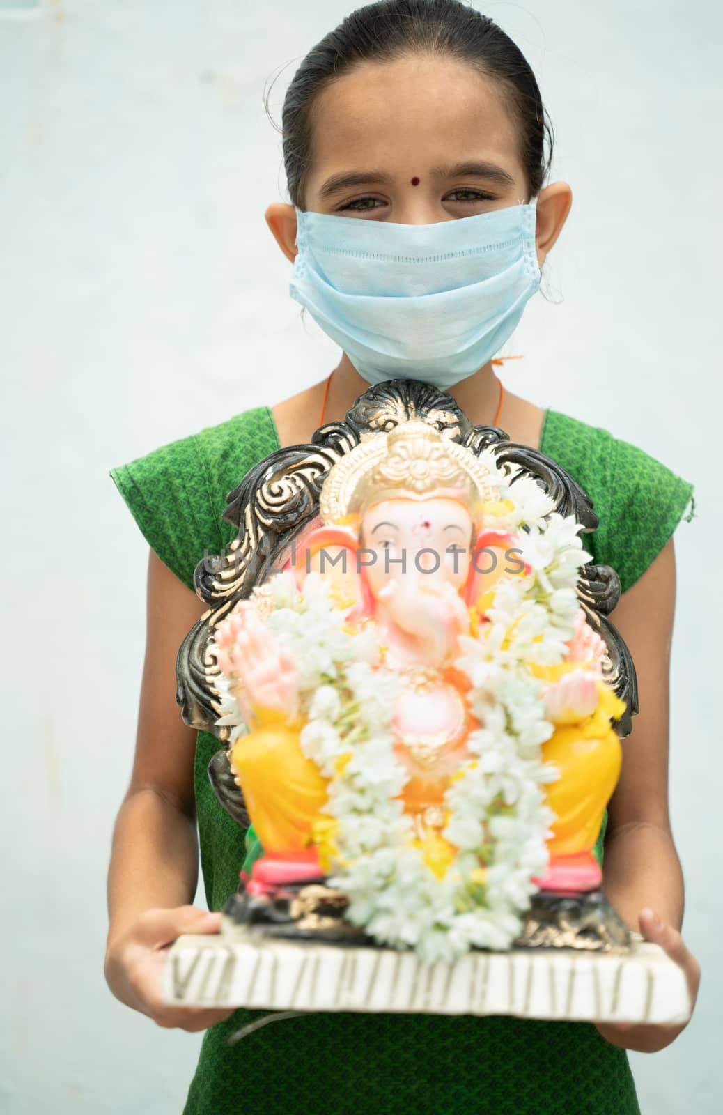 Girl Kid with medical mask holding Ganesha Idol on isolated background - concept of vinayaka Chaturthi festival celebrations during coronavirus or covid-19 pandemic by lakshmiprasad.maski@gmai.com