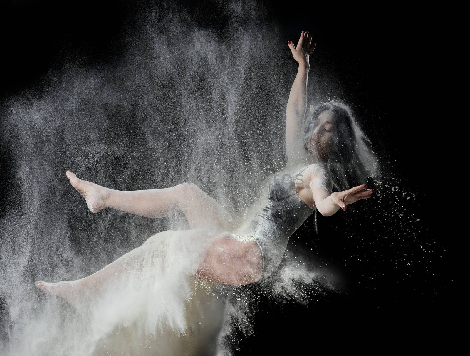 beautiful young caucasian woman in a black bodysuit with a sports figure soars on a black background in a spray of white flour, creative photo, levitation effect