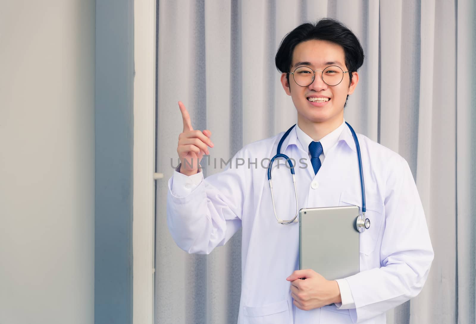 Portrait closeup of Happy Asian young doctor handsome man smiling in uniform and stethoscope neck strap holding smart digital tablet on hand and point finger to side away, healthcare medicine concept