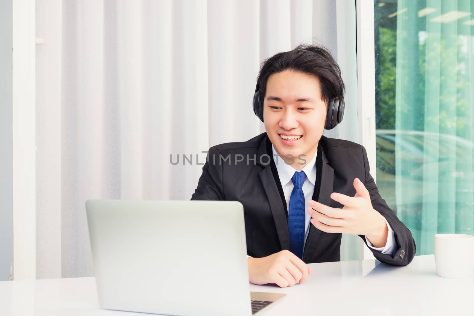 Work from home, Asian young businessman smile wearing headphones and suit video conference call or facetime on desk and raise his hand to explain to colleagues at home office