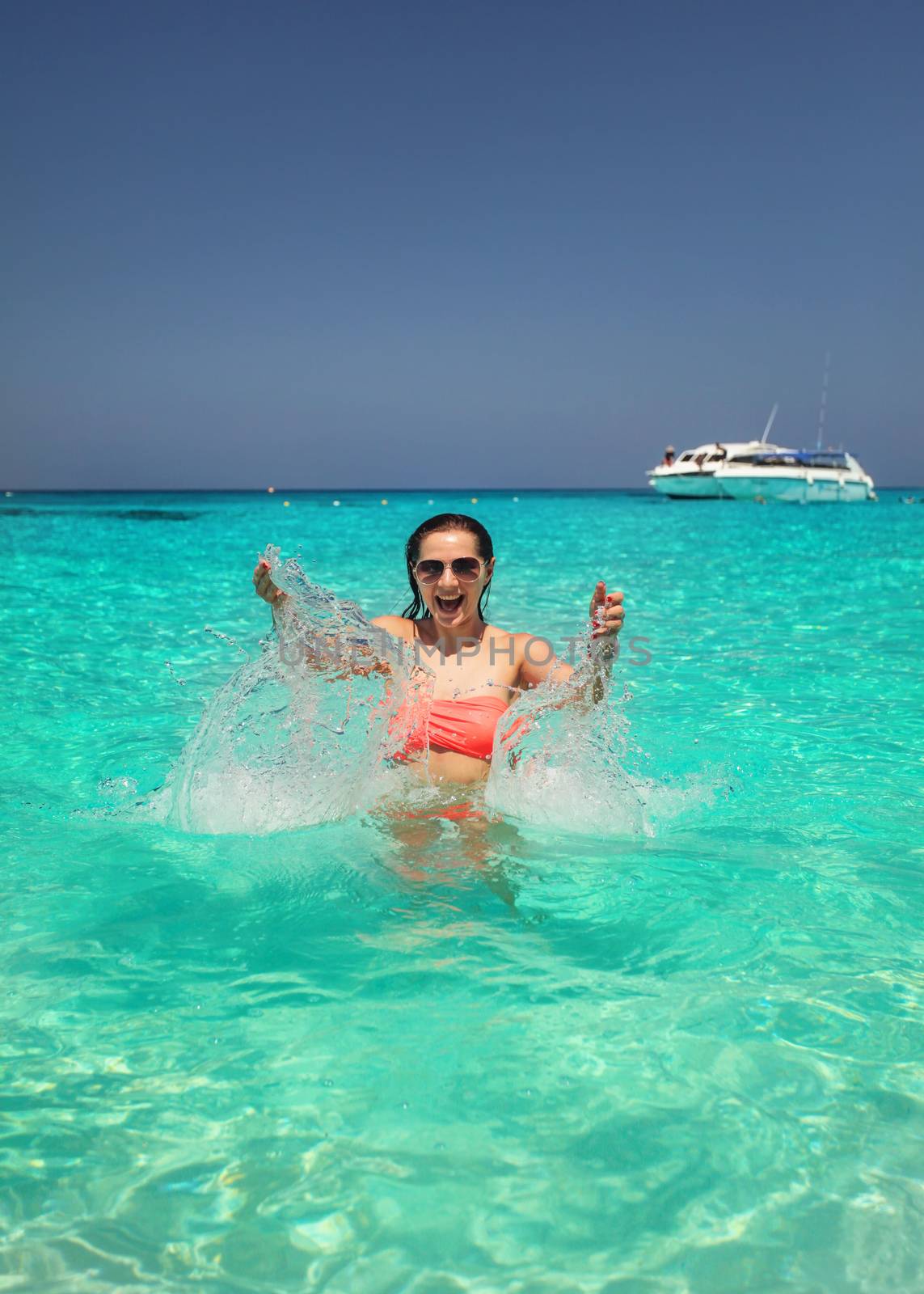 Sunglasses wearing young woman in crystal clear sea water splashing water in front of her with smiling happy face  expression. Similan Islands, Thailand