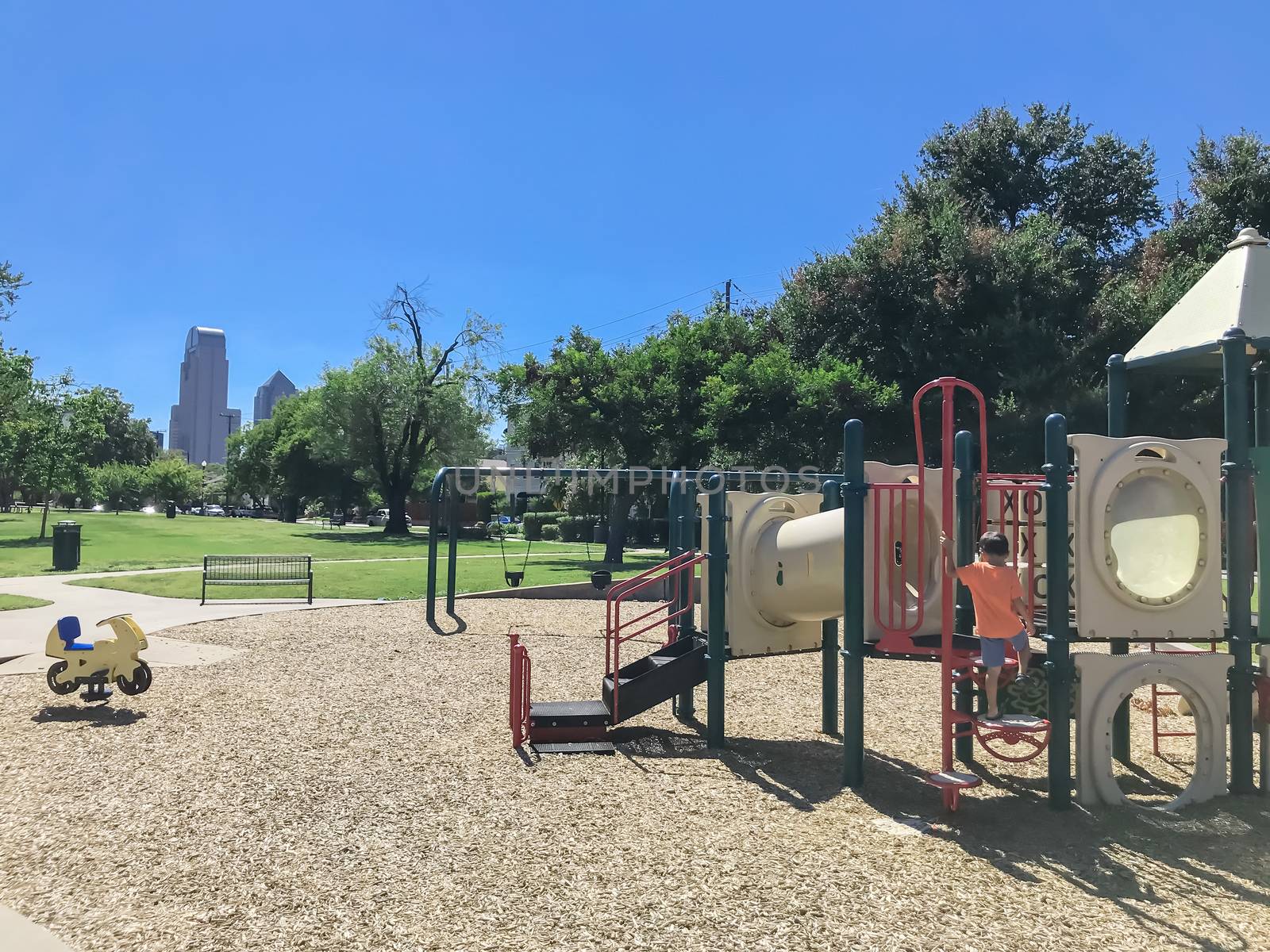 Kid playground with skylines background at public park surrounded by large trees in downtown Dallas, Texas, America. Empty recreation place in hot summer day with sunny clear blue sky.