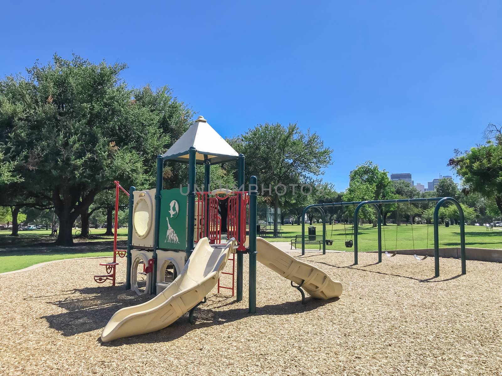 Colorful urban playground at public park surrounded by large trees in downtown Dallas, Texas, America. Empty recreation place in hot summer day with sunny clear blue sky.