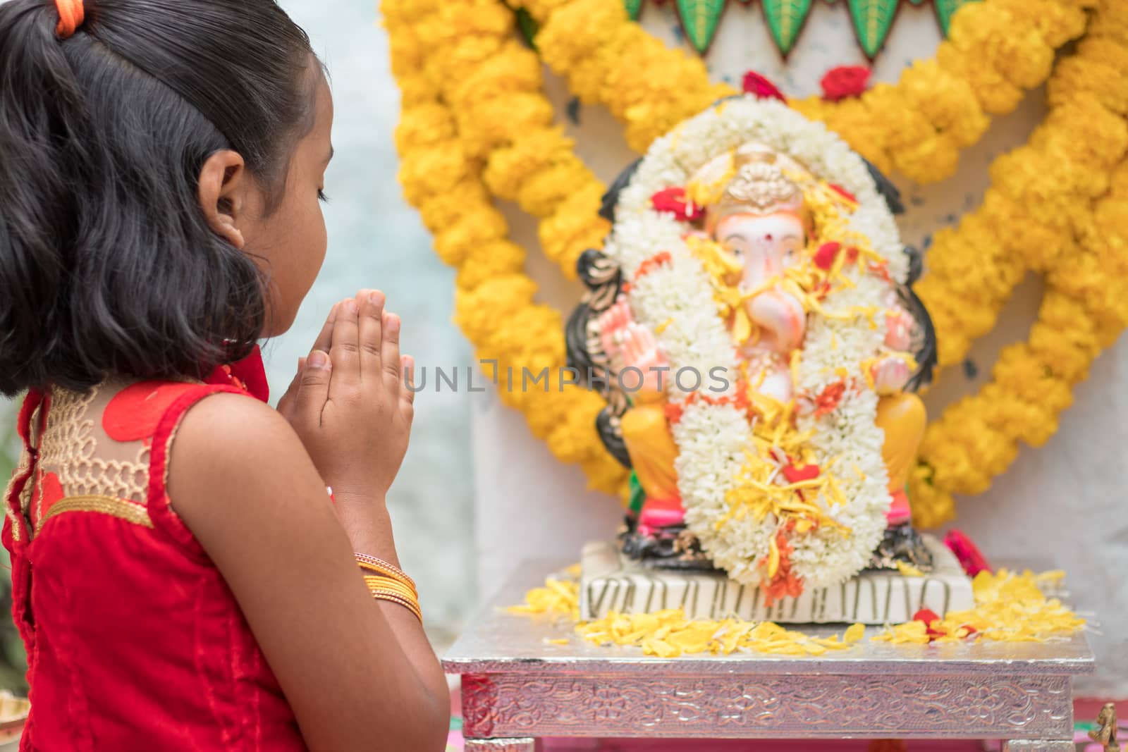 Girl Kid praying by closing eyes and folding hands in front of Lord Ganesha Idol during Ganesha or vinayaka Chaturthi festival ceremony at home - concept of Indian religious festival celebrations