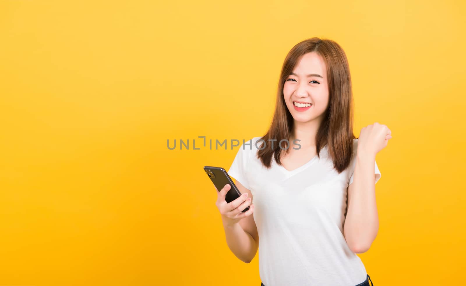 Asian happy portrait beautiful cute young woman teen smiling standing wear t-shirt celebrating win with smart mobile phone looking the phone isolated, studio shot on yellow background with copy space