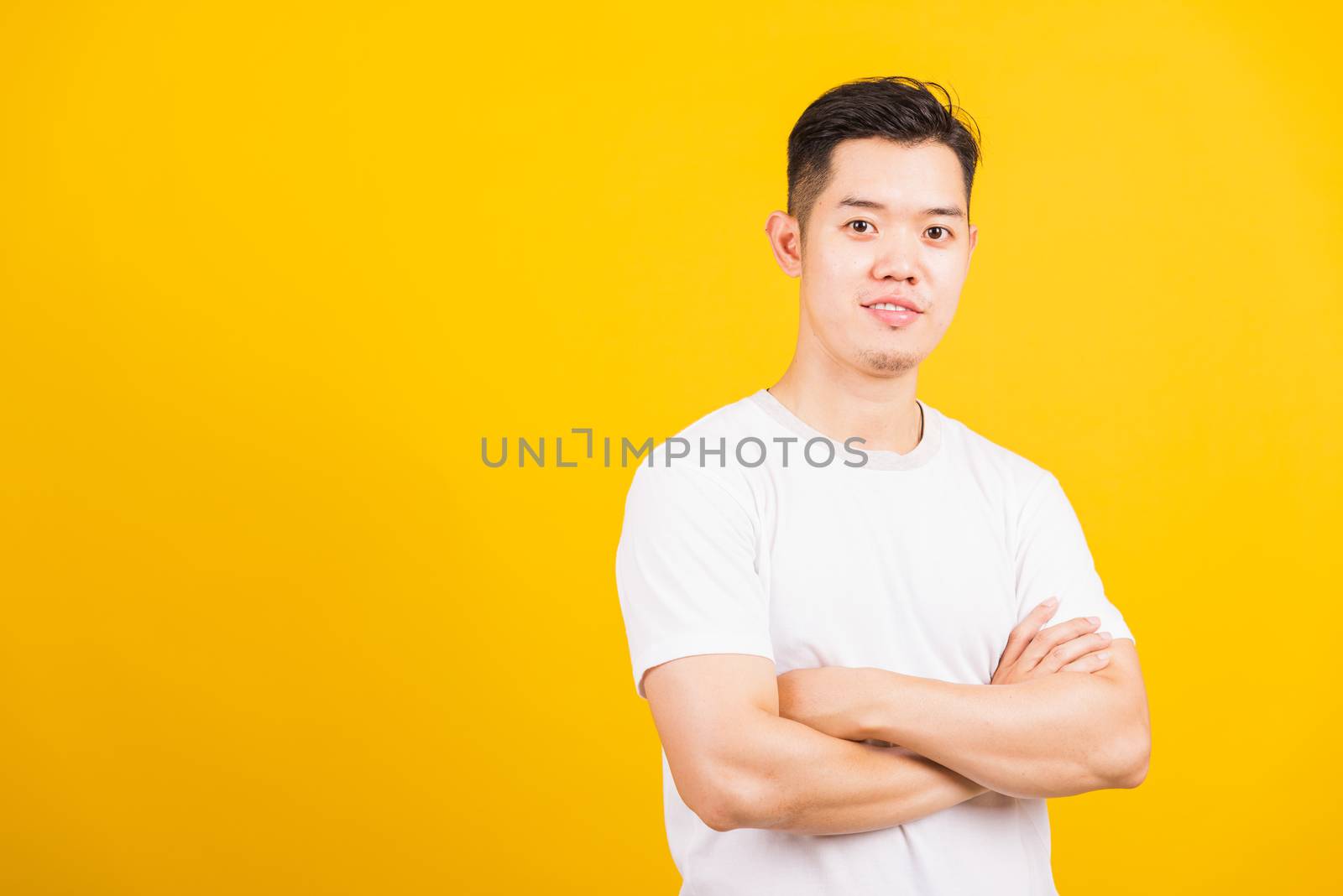 Portrait happy Asian handsome young man smiling standing wearing white t-shirt folded or crossed arm he looking to camera, studio shot isolated yellow background