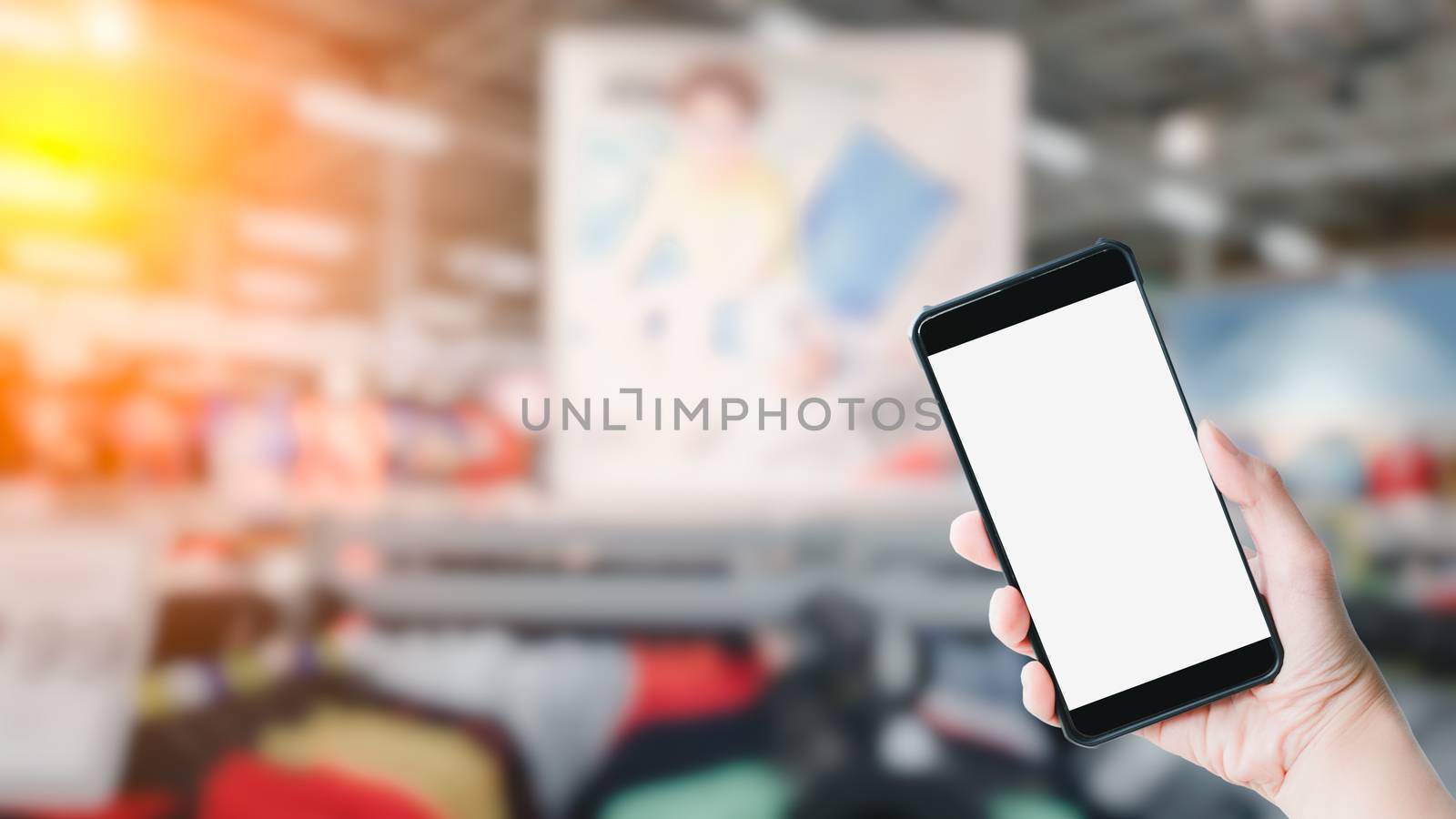 Shopping online concept. Hand holding black smartphone and white blank screen in blurry at shopping or department store background. Mock up shopping.
