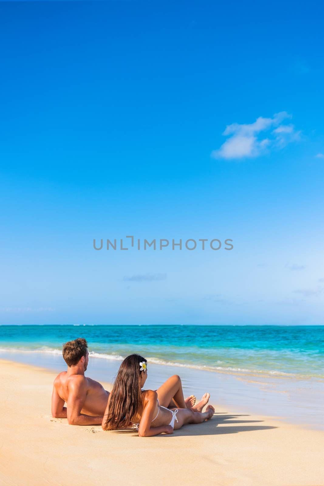 Beach vacation couple tanning under the tropical sun enjoying summer holidays traveling aroung the world. Young people lying down in paradise. Vertical background with copy space on sky.