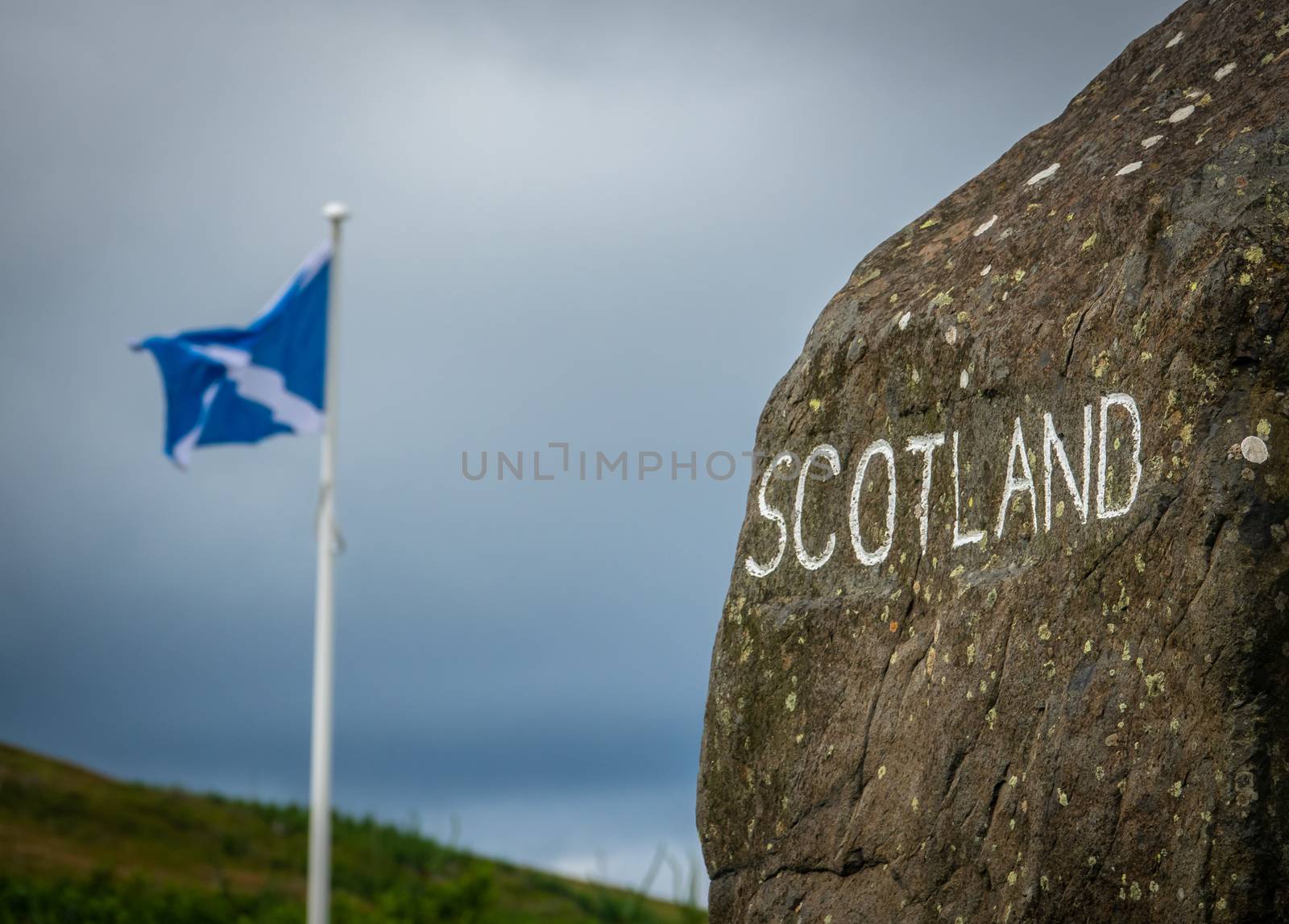 A Stone Sign At The Scottish Border With A Scotland Flag In The Distance