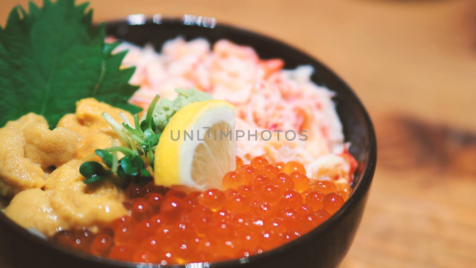 Close-up images of japanese seafood rice bowl or kaisendon sashimi donburi which have fresh ogura, uni, giant crab and topping with lemon on rice in the black bowl on the wood table from top view angle camera.